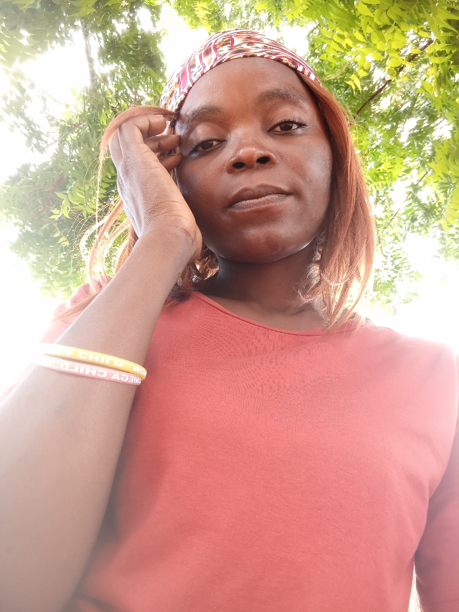 In Kason, Nigeria, a woman is photographed from a low angle, looking directly at the camera. She is wearing a red shirt and a patterned headscarf. Her right hand is raised to her forehead. Two bracelets are visible on her left wrist, one yellow and one pink, with text that is difficult to discern. The background consists of lush green foliage and bright sunlight filtering through the leaves.