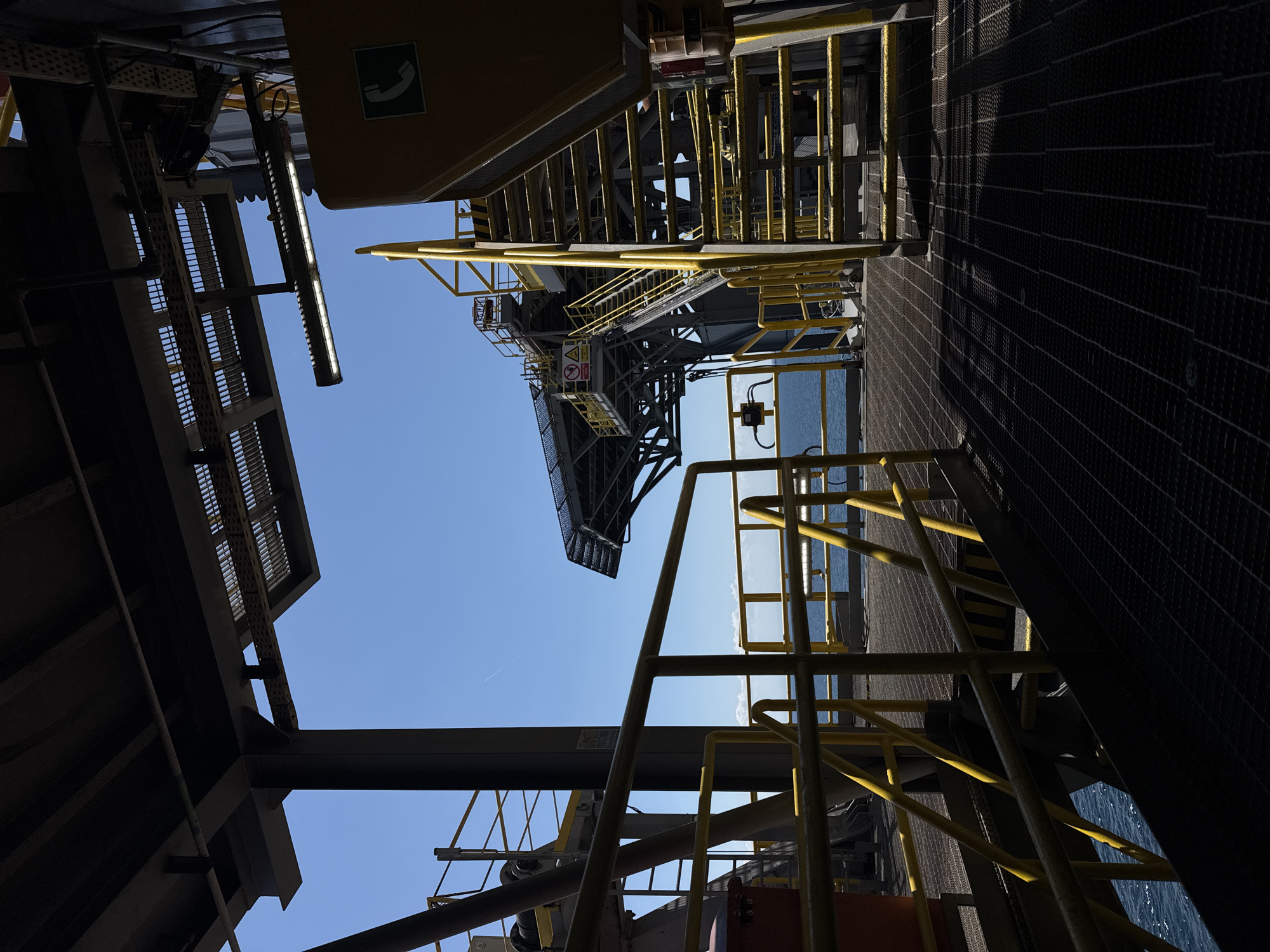 The image depicts the exterior of an industrial structure, likely an oil rig or offshore platform, under a clear blue sky. The foreground is dominated by dark, shadowed grating forming the walkway and a yellow metal railing. To the right, a bright yellow emergency phone booth and a set of metal stairs with yellow handrails are visible. In the midground, more yellow railings line a walkway that extends towards a large, complex metal structure with a helideck visible in the distance. The sea can be seen beyond the structure, with a few white clouds on the horizon. The lighting suggests it is daytime, with strong sunlight casting deep shadows. The location is identified as Unknown, Italy.