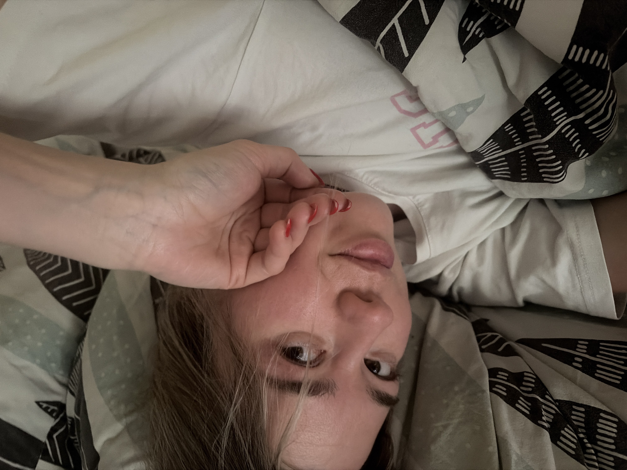 A woman lies in bed, her hand partially obscuring her face. Her fingernails are painted red. She wears a white T-shirt and is covered with a blanket that has a geometric black and white pattern. The background appears to be a bed. The city of Chaikovsky, Russia is not visually confirmed.