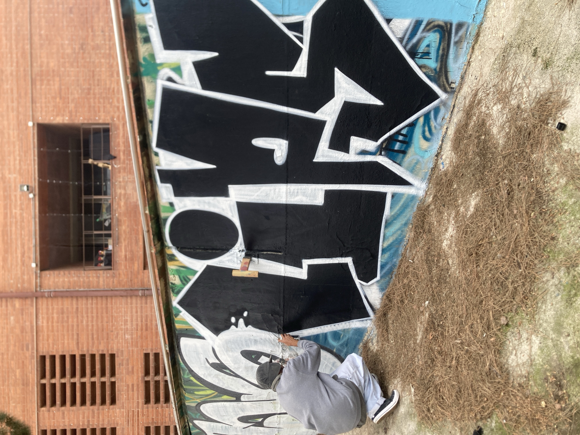 A person wearing a grey hoodie, light-colored pants, and black and white shoes is crouched down, painting a large piece of graffiti on a wall. The graffiti is primarily black and white with sharp, angular lines. The wall also features other graffiti beneath the new artwork, including blue and green elements. The wall is adjacent to a patch of dry grass and dirt. To the left, there is a brick building with a window and vertical vents. The presence of the brick building style suggests that this photograph was taken at the Parc del Fòrum in Barcelona, Spain.