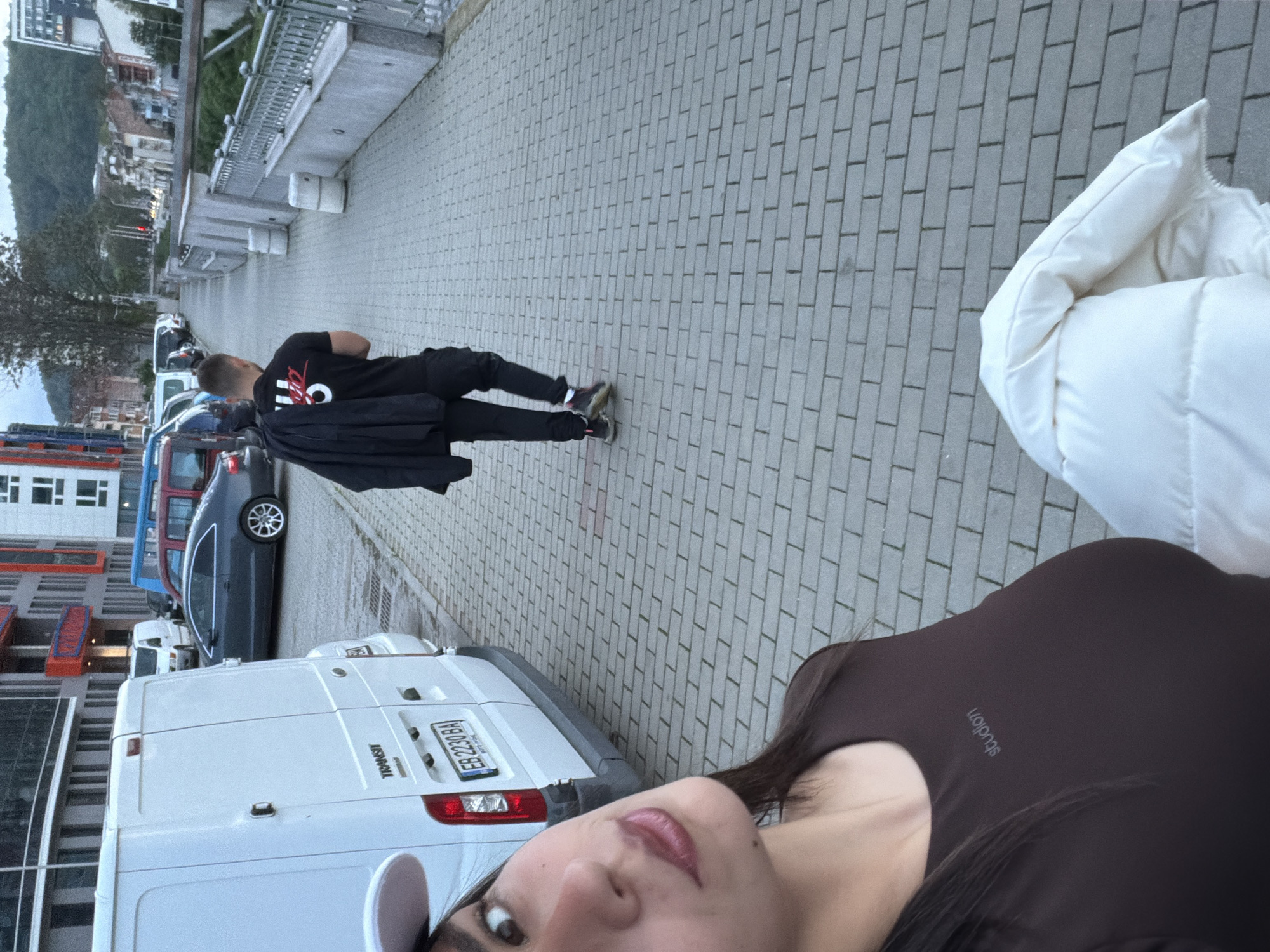 The image depicts a scene on a paved street in Gabrovo, Bulgaria. A woman is taking a selfie. She is wearing a brown top and a white baseball cap. Part of a white puffy jacket is visible near her. Behind her is a white van, and its license plate number is EB 2330 BA. A man is walking on the sidewalk near parked cars and a grey railing. In the background is a building with red details. In the distance, a tree-covered hill is visible.