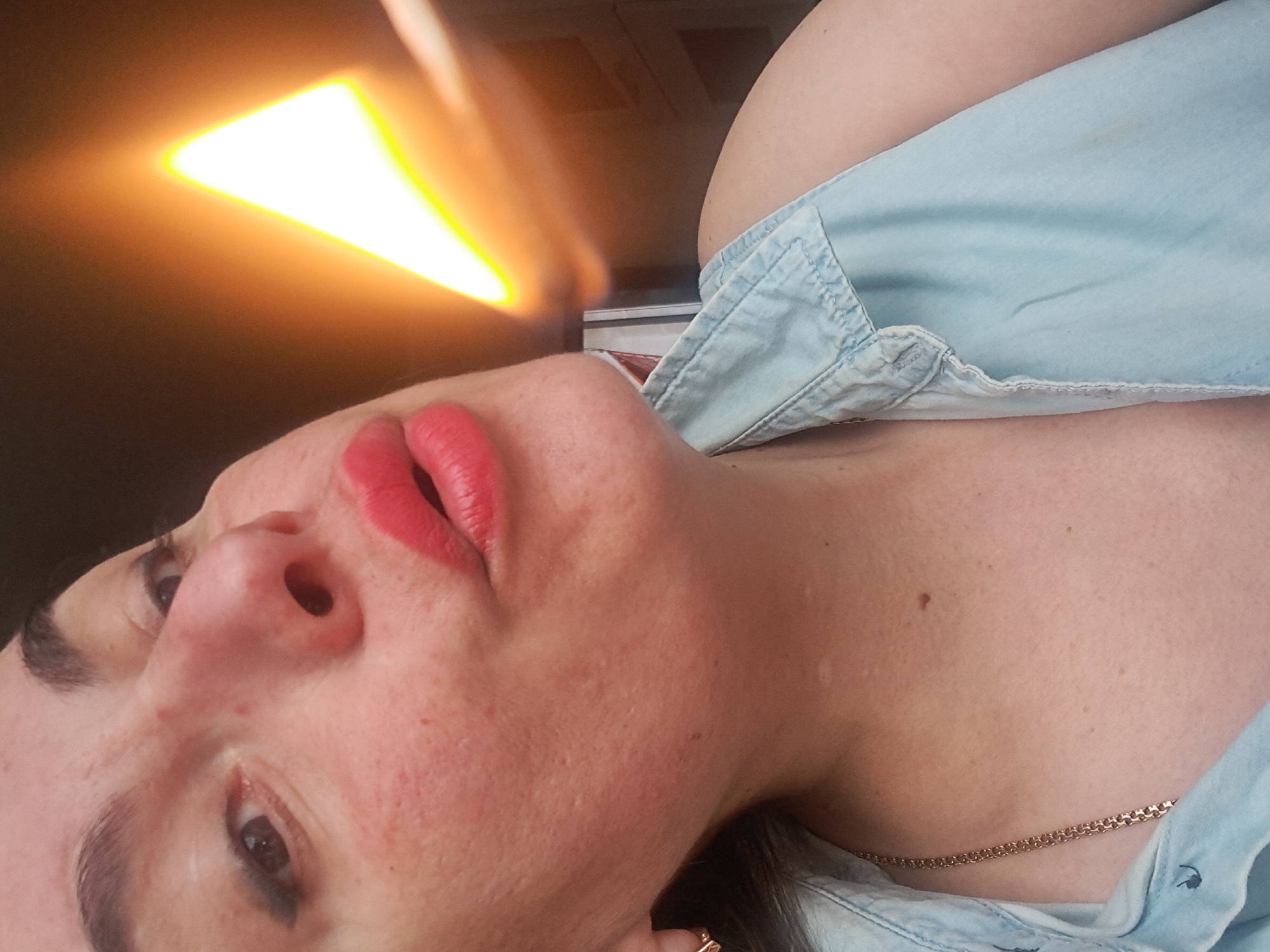 A close-up, slightly rotated photograph shows a woman's face and upper torso. She has fair skin, dark eyebrows, and brown eyes. Her lips are painted a bright red. She is wearing a light blue denim shirt and a gold chain necklace is visible. A bright, blurred triangle of light is present in the upper left of the image. The setting appears to be indoors, possibly in a vehicle, based on the light source and background, but there is no specific evidence to indicate it is in Elektrostal, Russia.