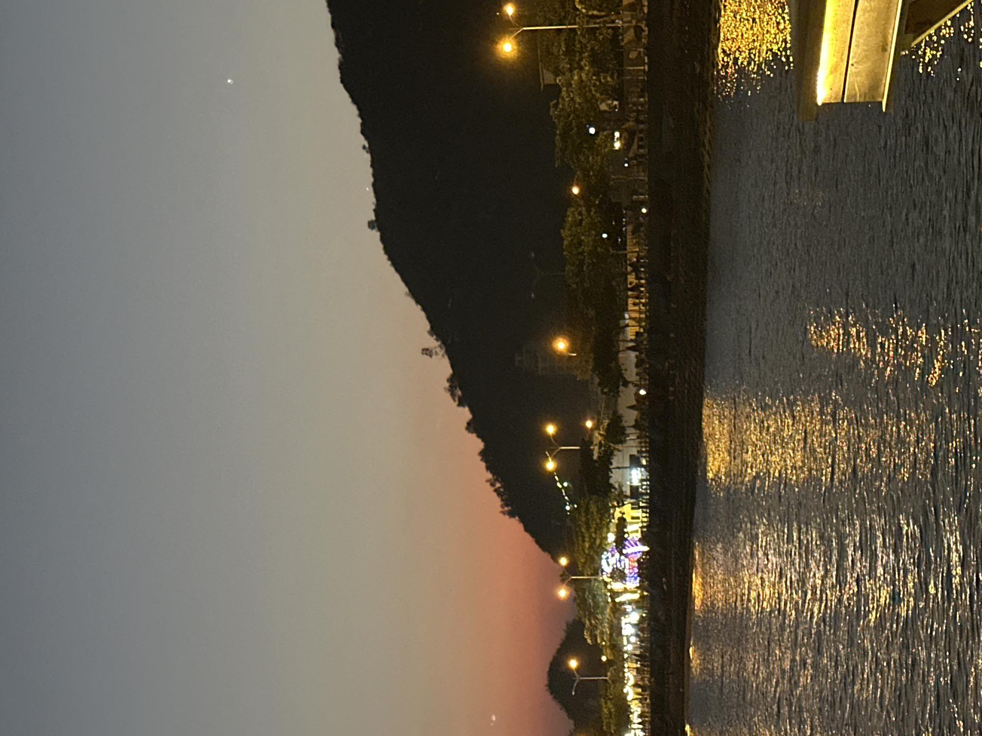 The image displays an evening scene in a coastal or bay area of Nha Trang, Vietnam, during twilight or early night. The sky transitions from dark grey-blue overhead to an orange-red glow near the horizon. A single bright celestial body is visible in the upper right quadrant of the sky.
The foreground is dominated by a dark body of water, which reflects numerous artificial light sources as shimmering golden-orange streaks. A dark-colored structure with a flat, illuminated top edge, possibly a pier or boat element, extends into the lower right corner of the frame.
The midground features an illuminated urban waterfront. Multiple tall streetlights with yellow-orange light sources are prominently visible, casting reflections onto the water. Trees line this area, some adorned with decorative, multicolored lights. A large structure, heavily embellished with various colored lights, stands centrally on the promenade. Indistinguishable silhouettes of individuals are present on the promenade, indicating pedestrian activity.
The background is occupied by a large, dark, tree-covered hill or mountain, silhouetted against the sky. Faint, scattered lights are discernible on its slopes, suggesting minimal development or infrastructure.