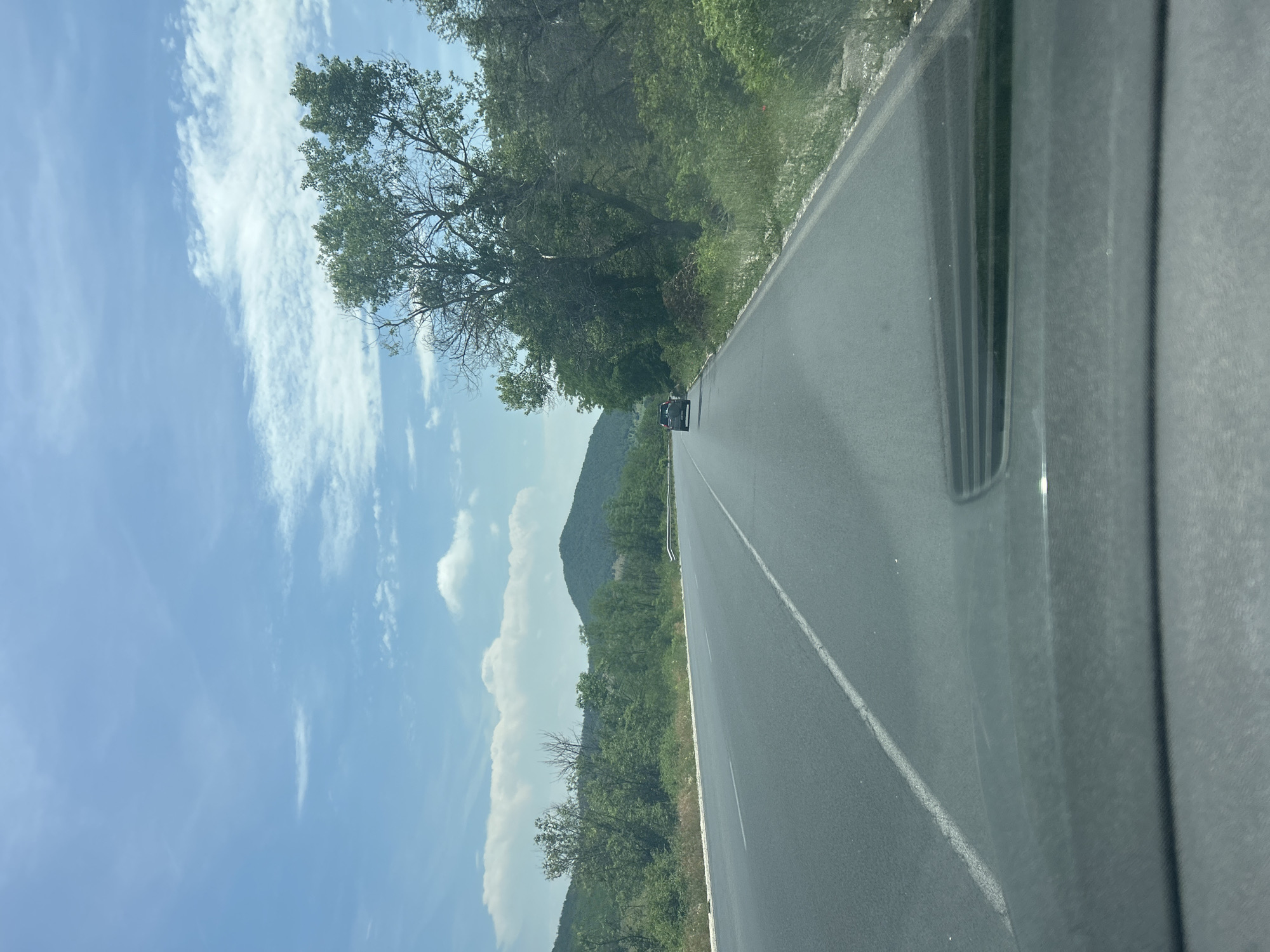 A vertical photograph captures a road journey through a scenic landscape in Mihiltsi, Bulgaria, viewed from inside a vehicle. The image depicts a two-lane asphalt road extending into the distance, marked by a solid white line on the right shoulder and a dashed white line separating the lanes. Lush, dense green trees and bushes line both sides of the road, creating a natural, verdant tunnel.
In the background, a range of rolling green mountains or large hills rises under a bright, clear blue sky adorned with scattered white, wispy clouds. A dark-colored car is visible far down the road, heading away from the viewer. The right edge of the frame shows a partial view of the interior of the vehicle from which the photo was taken, including a dark dashboard or door panel and an air vent, suggesting the photo was captured during a car trip.
The time of day appears to be sunny daytime, with clear weather conditions. The overall scene conveys a sense of serene travel through a natural, mountainous region. There is no visible text in the image.