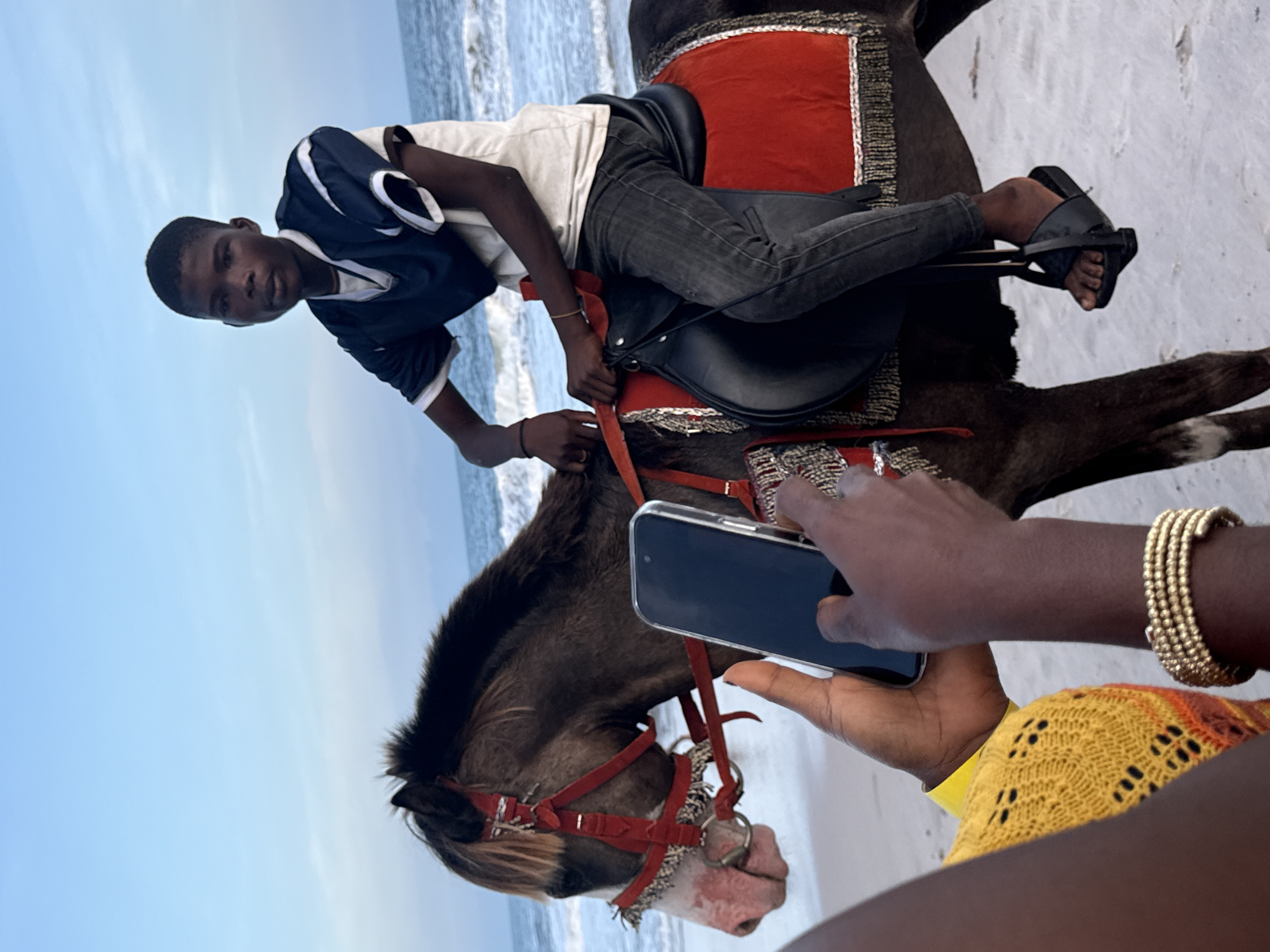 A photograph taken in Mopo Onijebu, Nigeria, depicts a young person riding a horse on a beach. The rider, wearing a white t-shirt, dark-colored pants, and a dark jacket with white trim, is positioned atop a dark-colored horse adorned with a red and silver saddle and bridle. The horse stands on the sandy beach, with the ocean and sky visible in the background. In the foreground, a person holds a smartphone, likely taking a photo of the rider. The photographer is wearing a yellow and orange patterned top and has gold bracelets on their wrist. The scene seems to be a tourist activity, with someone capturing the moment of the horseback ride.