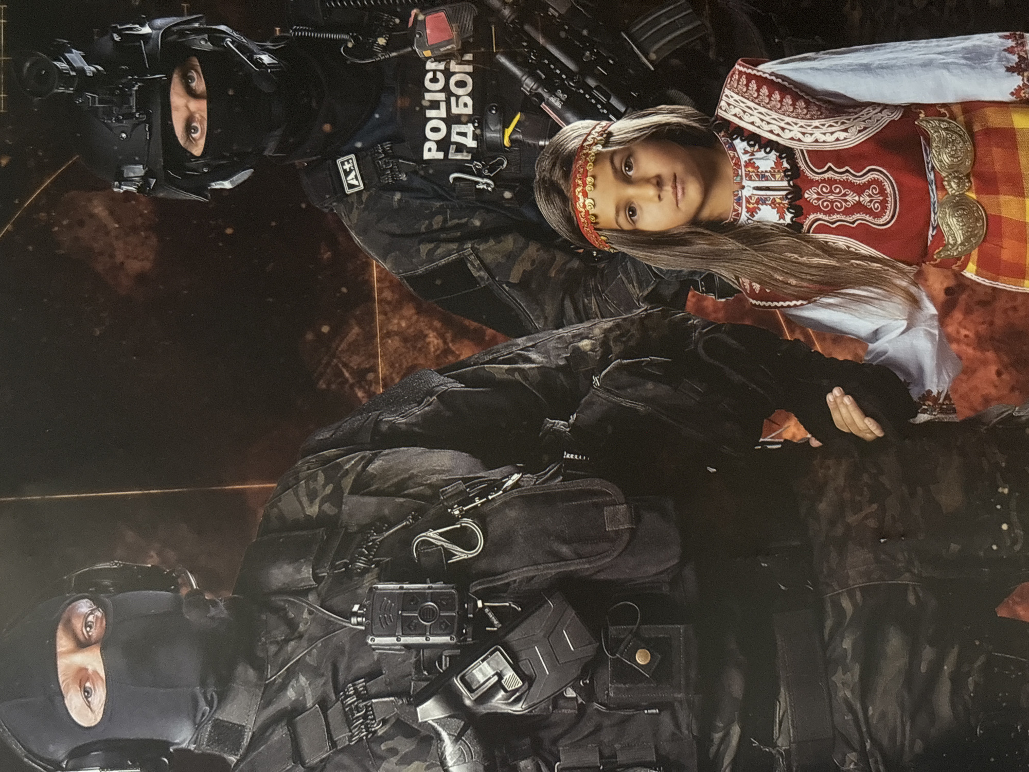 The image depicts a superimposed scene with a juxtaposition of law enforcement and civilian elements, potentially related to Plovdiv, Bulgaria.
On the upper half, a tactical officer in full gear, including a helmet with night vision equipment, is positioned. The officer wears a vest labeled "POLICE ГДБОП," indicating affiliation with the General Directorate for Combating Organized Crime in Bulgaria. Beside the officer, a young girl wearing traditional Bulgarian folk attire is partially visible, specifically a red and white embroidered top and an orange and yellow plaid skirt, along with a decorative headband.
In the lower half of the image, another tactical officer is seen in similar black tactical gear. The background features a dark, textured surface, possibly suggesting a digitally altered or composite image.
The image suggests a symbolic or thematic representation, with the juxtaposition of heavily armed tactical units and a child in traditional clothing. There is no specific location visible, but considering the "POLICE ГДБОП" designation and the traditional clothing, it is possible the image alludes to themes or issues related to Bulgaria. The setting lacks discernible features to specifically associate it with Plovdiv.