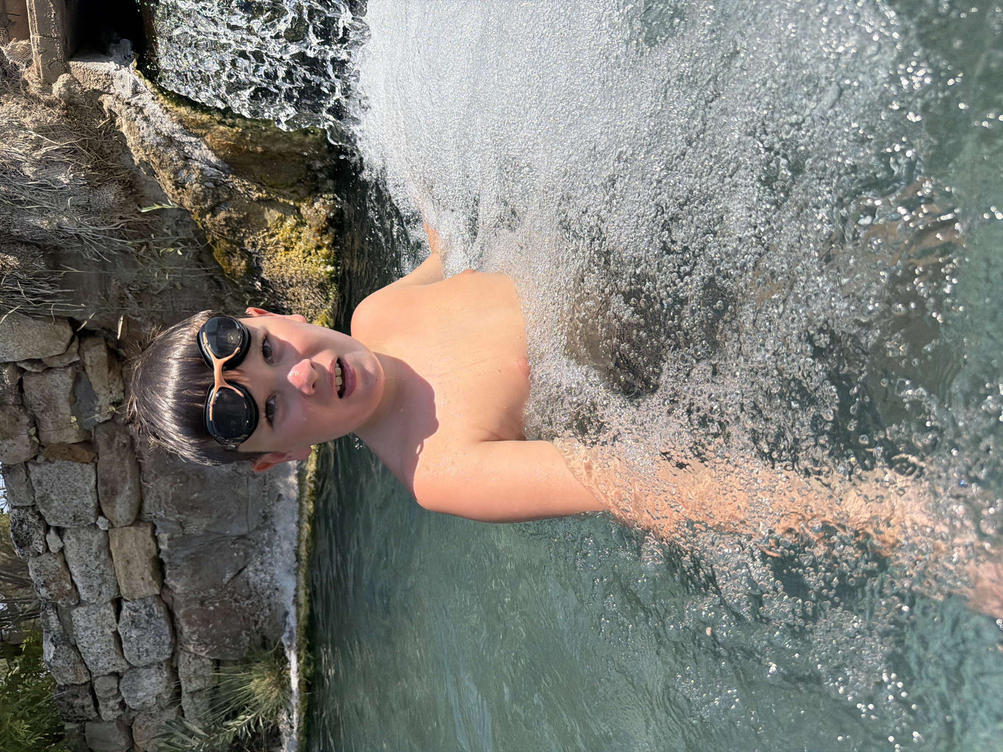 This is a medium shot, portrait-oriented photograph of a young boy in a pool of clear, turquoise water, with a waterfall cascading in the background. The boy is wearing black and orange swimming goggles and is looking at the camera with a playful expression, his mouth slightly open. He is shirtless and his skin is flushed, suggesting he has been in the water for a while.
The setting appears to be a natural hot spring, given the context of Pamukkale, Turkey, known for its travertine terraces and thermal waters. The water in the pool is calm and reflective, with a gentle ripple. The waterfall behind the boy is frothy and white, creating a dynamic contrast with the still water in the foreground. The background also shows some rocky, textured elements of the natural landscape.
The lighting suggests it is daytime, with the sun casting bright highlights on the water and the boy's skin, indicating a sunny day. The overall mood of the photograph is one of joy and recreation, capturing a moment of carefree fun in a beautiful natural setting. There is no visible text in the image.