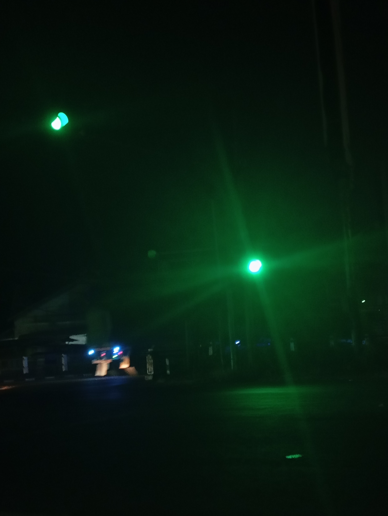The image depicts a nighttime street scene in Sokaraja, Indonesia, dominated by green light emanating from traffic signals and other illuminated sources. In the upper left quadrant, a green traffic light is visible, casting a diffused glow. To the right, another prominent green light source, likely a traffic signal or street lamp, creates strong light beams that extend across the frame. In the lower portion of the image, a blurred vehicle with blue and red lights is partially visible, suggesting it may be stopped or moving slowly. Structures, possibly buildings or road infrastructure, are discernible as dark silhouettes in the background. The overall environment is dark and atmospheric, with the green light providing the primary illumination and lending a somewhat surreal quality to the scene. There are no people clearly visible in the image.