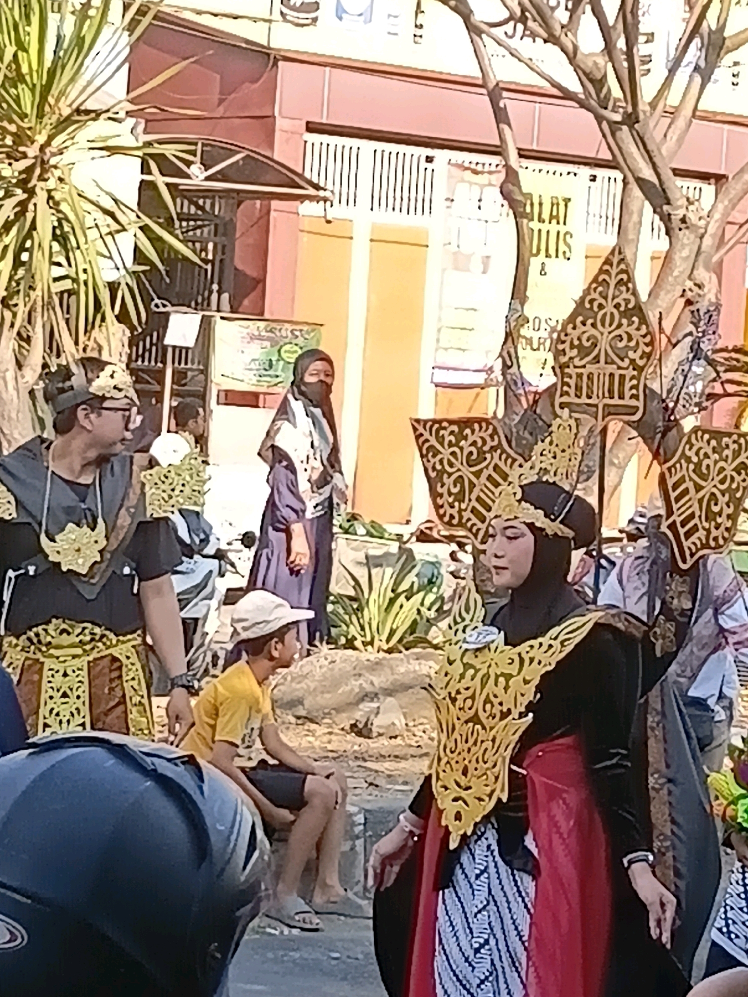 This image captures a street scene in Wadungasri, Indonesia, possibly during a cultural event. In the foreground, a person in elaborate traditional attire with a black hijab and a golden headdress is looking to their left. The attire includes a black top with a flowing red skirt and a patterned sarong-like lower garment, adorned with ornate gold accents on the chest and shoulders. To the left of this individual, another person, appearing to be male, is also wearing traditional attire in black and gold. He is wearing a headpiece and glasses, and his gaze is directed to his left. A young boy in a yellow t-shirt and shorts sits on a low wall in the middle ground, looking to his right. In the background, a woman in a dark hijab and long purple garment stands, facing forward. Other individuals are visible further back. Behind the people, a building with a pink facade and barred windows is partially obscured by trees. Banners with Indonesian text are also visible on the building. The ground appears to be a paved street with a curb. The overall atmosphere suggests a public gathering or parade.