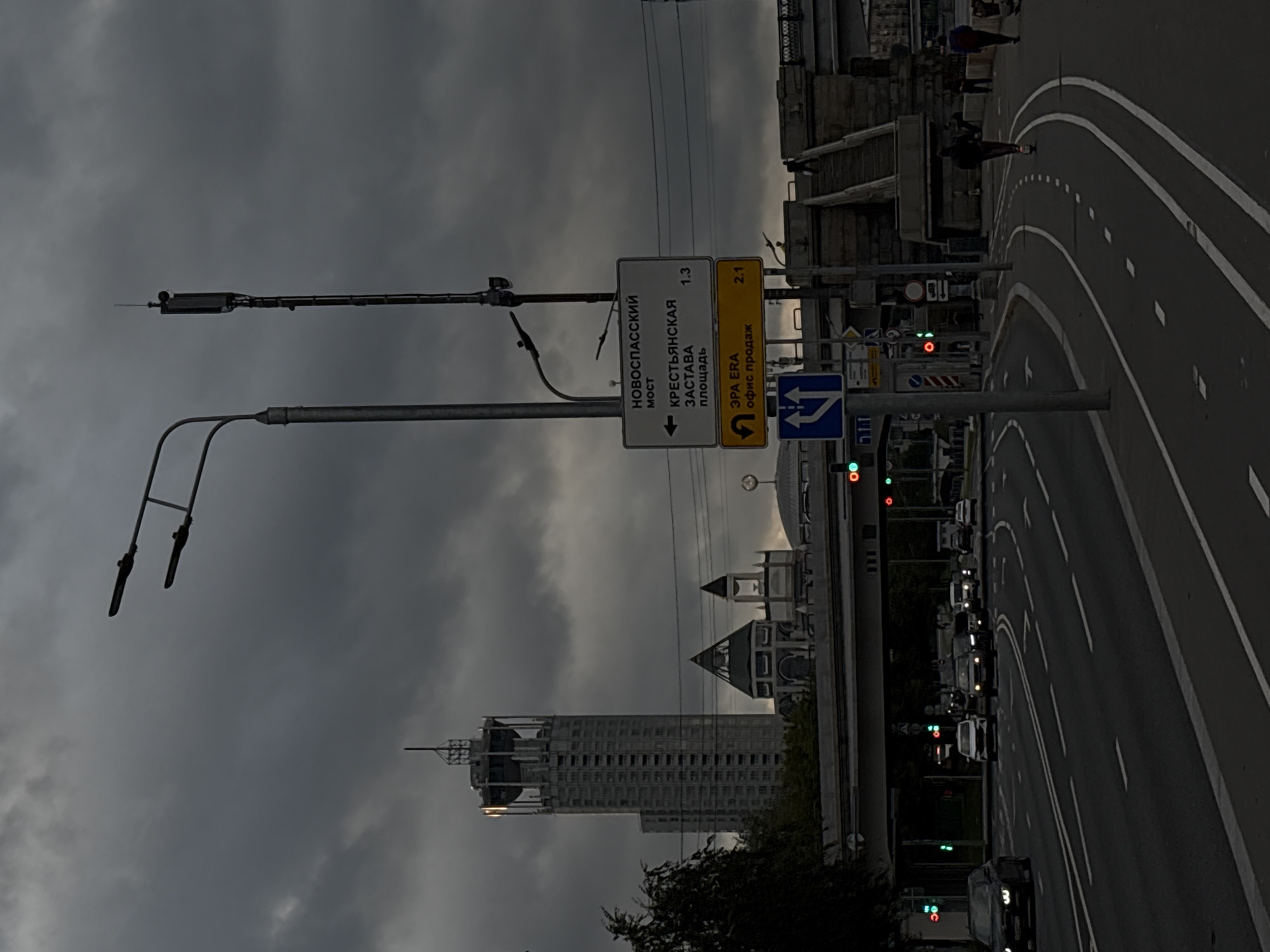 This is a street-level view of a road in Moscow, Russia, on a cloudy day. In the background, a tall, modern skyscraper with a distinctive observation deck at the top dominates the skyline. Below and to the right of the skyscraper are several distinctive, dome-shaped buildings, possibly religious or cultural structures.
A street signpost is prominently placed, displaying directions in Russian. The upper sign indicates "НОВОСПАССКИЙ МОСТ" (Novospassky Bridge) with an arrow pointing left, and "КРЕСТЬЯНСКАЯ ЗАСТАВА ПЛОЩАДЬ" (Krestyanskaya Zastava Square) also with an arrow pointing left, accompanied by "1.3". The lower yellow sign reads "ЭРА ERA" and "офис продаж" (sales office), with an arrow indicating a right turn, followed by "2.1".
Several cars are visible on the multi-lane road, some stopped at traffic lights, while others are moving. Traffic lights show green for some lanes. There is a bike lane marked with white dashed lines alongside the road.
In the foreground, a person is riding an electric scooter on the bike lane, heading away from the camera. Further back, two other individuals are walking on the sidewalk or path. A cell tower with antennas is mounted on a pole near the street sign. Overhead, a street lamp with two lights is visible against the dark, cloudy sky. The overall atmosphere is somber due to the overcast conditions.