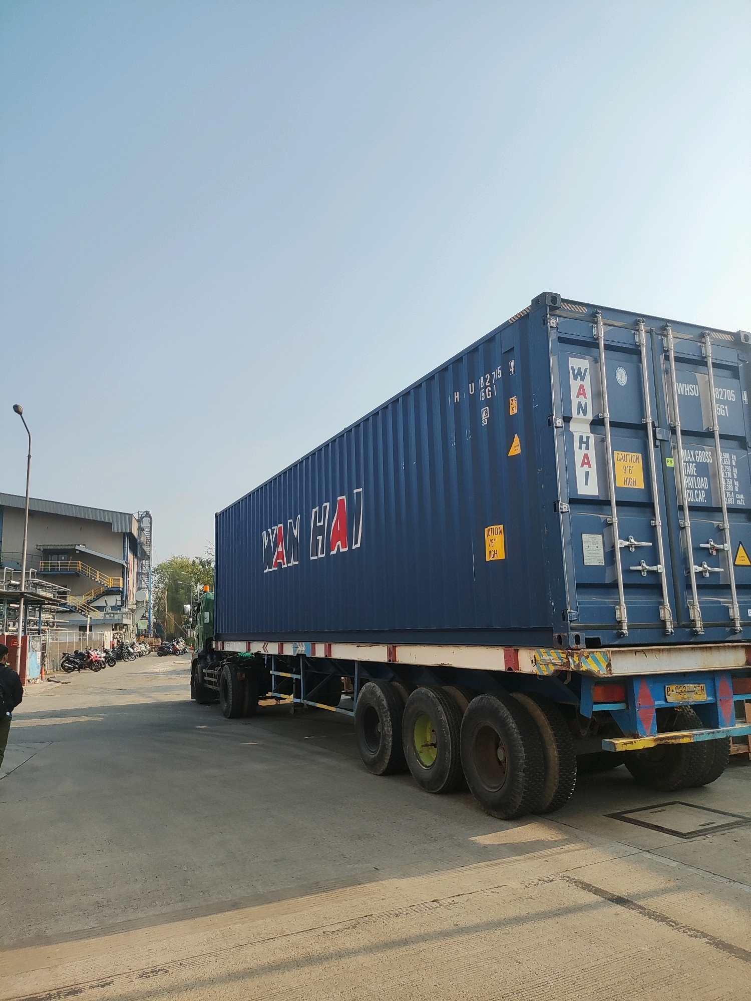 A blue shipping container, marked with "WAN HAI" and identification numbers like "WHSU 8275", is loaded onto a truck. The truck, with multiple axles and large tires, is parked on a paved surface. In the background, a building with a grey facade and yellow stairs is visible, along with a row of parked motorcycles. A lamppost stands to the left of the scene. The sky is clear and blue. The license plate on the truck appears to be "B 9339 KEM". The image was taken in East Jakarta, Indonesia.
