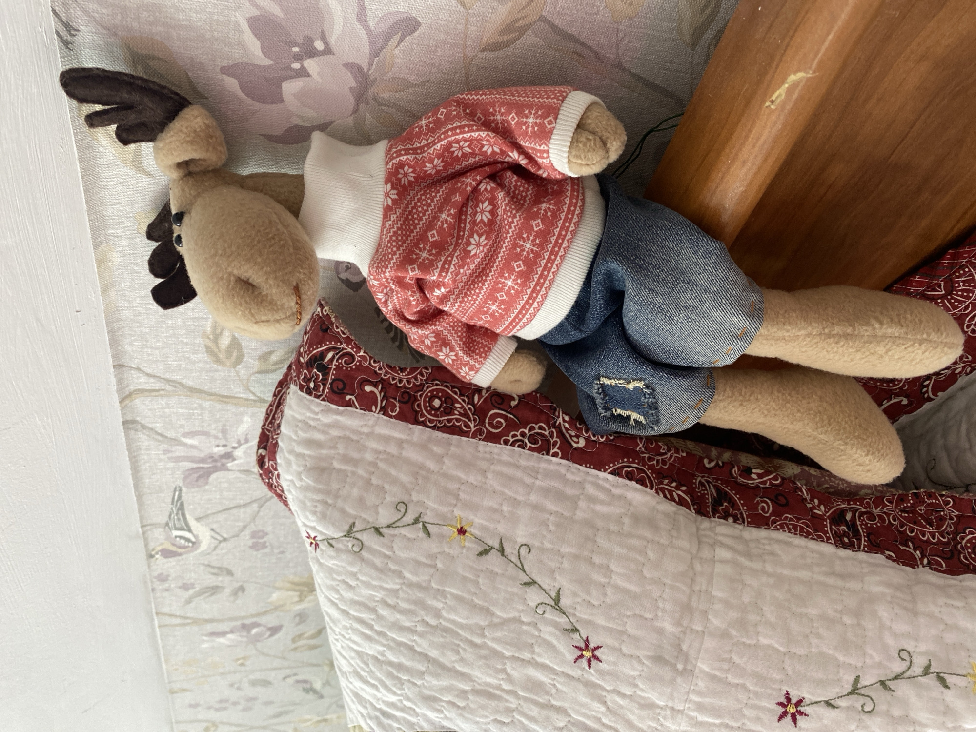 A plush deer toy is centrally featured in this image, lying on a bed or soft surface. The toy has a light tan body, dark brown felt antlers, and small black eyes. It is dressed in a white long-sleeved undershirt, a red sweater adorned with a white Fair Isle pattern featuring snowflakes and geometric shapes, and blue denim shorts with distressed patches on the thighs. The toy's limbs are soft and plush.
The deer is resting on a white quilted pillow or blanket, which has a wavy stitched pattern and delicate embroidered floral vines with small yellow and reddish-pink flowers. This white quilted section is bordered by a wider band of fabric featuring a red and white paisley pattern.
In the background, a light-colored wallpaper or fabric with a muted floral design in shades of purple, green, and brown is visible. A small bird motif can also be seen within this pattern. To the right of the toy, a portion of a wooden furniture piece, possibly a headboard or bed frame, is visible, showing a light brown wood grain and a minor scratch.
The setting appears to be an indoor space, likely a bedroom or living room. There is no specific activity taking place; the toy is simply posed or resting. The lighting suggests ambient indoor light. No text is visible in the image.