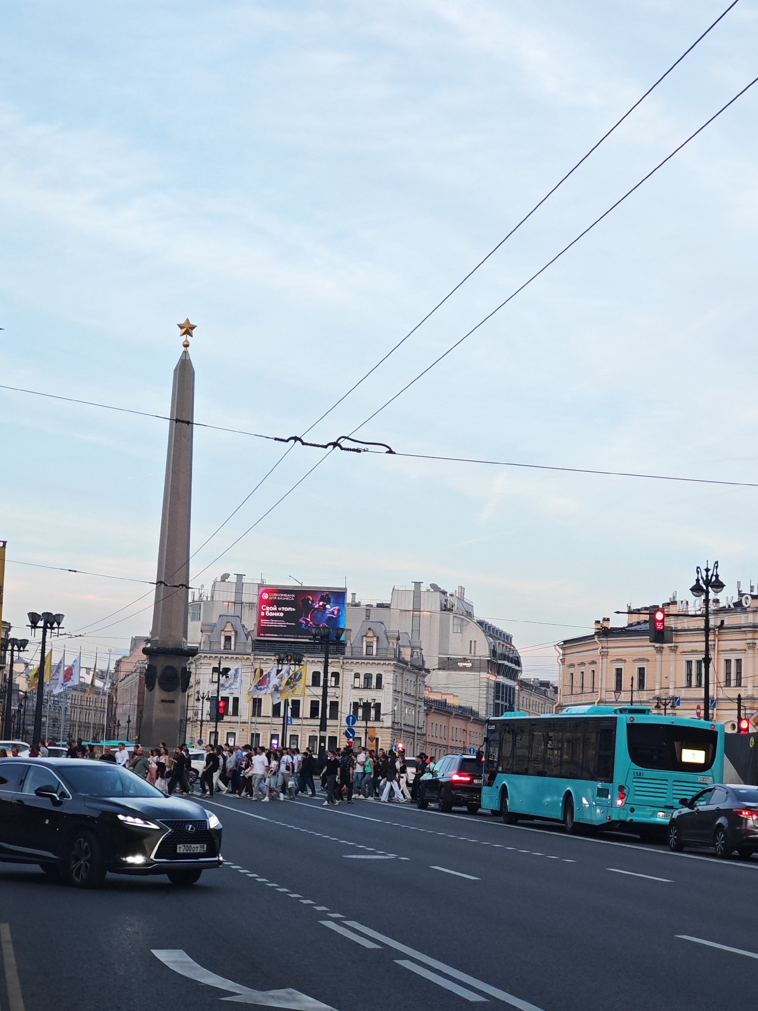 The image presents an urban scene at Vosstaniya Square in Saint Petersburg, Russia, observed during what appears to be late afternoon or early evening. The foreground features an asphalt road with white lane markings, including solid lines, dashed lines, and a right-turn arrow. A black Lexus RX SUV with illuminated headlights and a Russian license plate "T700OT 98" is positioned on the left side of the road, facing left.
In the midground, a broad pedestrian crossing is occupied by a large group of people walking from the left to the right side of the street. To the left of the pedestrians and central to the image stands the Obelisk of Hero City Leningrad, a tall, grey, tapering stone column topped with a golden five-pointed star. To the right of the pedestrian crossing, a light blue city bus (number 1581, route 7) is stationary in the right lane, facing away from the viewer. Behind the bus, a dark sedan is also stopped. A red traffic light, displaying a countdown of "55" seconds, is visible on a pole to the far right.
The background consists of multi-story buildings with classical architectural elements. One building behind the obelisk features a large digital billboard displaying colorful advertisements, some with Russian text. Several yellow and white flags are suspended from poles along the buildings near the obelisk. Overhead, multiple dark power lines traverse the pale blue sky, which contains faint, wispy cloud formations.
