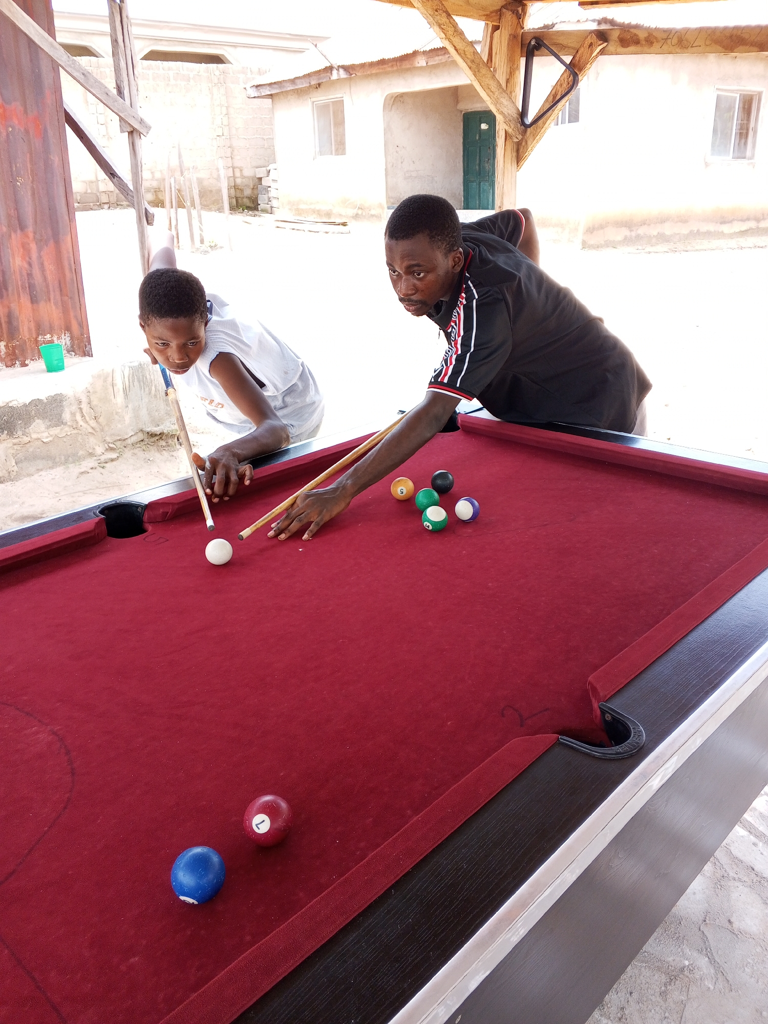 Two young Black individuals are playing pool on a maroon felt-covered pool table outdoors, under a wooden shelter with a corrugated metal roof. The person on the left, wearing a white tank top, is leaning over the table, aiming the cue stick towards a white cue ball. They appear focused on the shot. The person on the right, wearing a black t-shirt with red and white striped trim on the sleeves, is also engaged in the game, holding a cue stick. Several billiard balls are scattered on the table: a solid blue ball and a striped red ball with the number '1' are on the lower part of the table, while a solid yellow ball, a solid black ball, a striped green ball with the number '5', and a striped purple ball with the number '14' are clustered in the middle. The setting appears to be a casual outdoor area, possibly a backyard or a communal space, with concrete ground and simple buildings in the background under a bright, sunny sky, suggesting it is daytime. The overall mood seems relaxed and recreational. The scene is set in Lokoja, Nigeria.