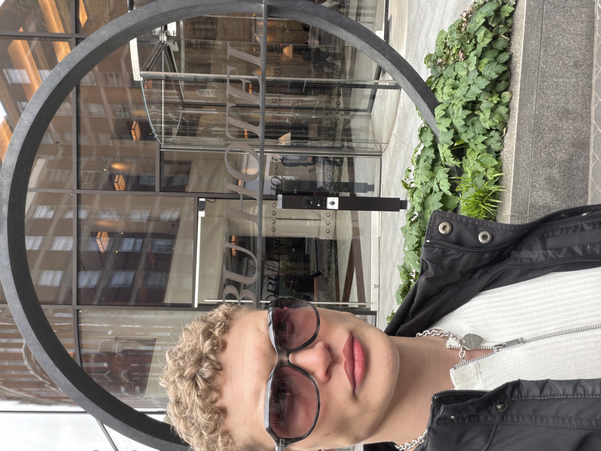 A young individual with fair skin and curly blonde hair is positioned in the foreground of the image. They are wearing dark sunglasses, a white ribbed top with a zipper, a silver chain necklace with a heart-shaped pendant, and a black jacket.
Behind the individual is a modern building with a large circular metal architectural feature in front of the entrance. The building appears to be a hotel, as the word "Hotel" is visible through the circular frame. The entrance includes glass doors and windows. The building's design reflects other buildings in the vicinity.
To the right of the circular frame, there is a patch of green ivy or similar climbing plants. The setting appears to be a city street. The overall scene suggests a location in an urban environment with modern architecture.
The architectural style and visible signage indicate this is the entrance to the Bvlgari Hotel London in Knightsbridge, London, United Kingdom.
