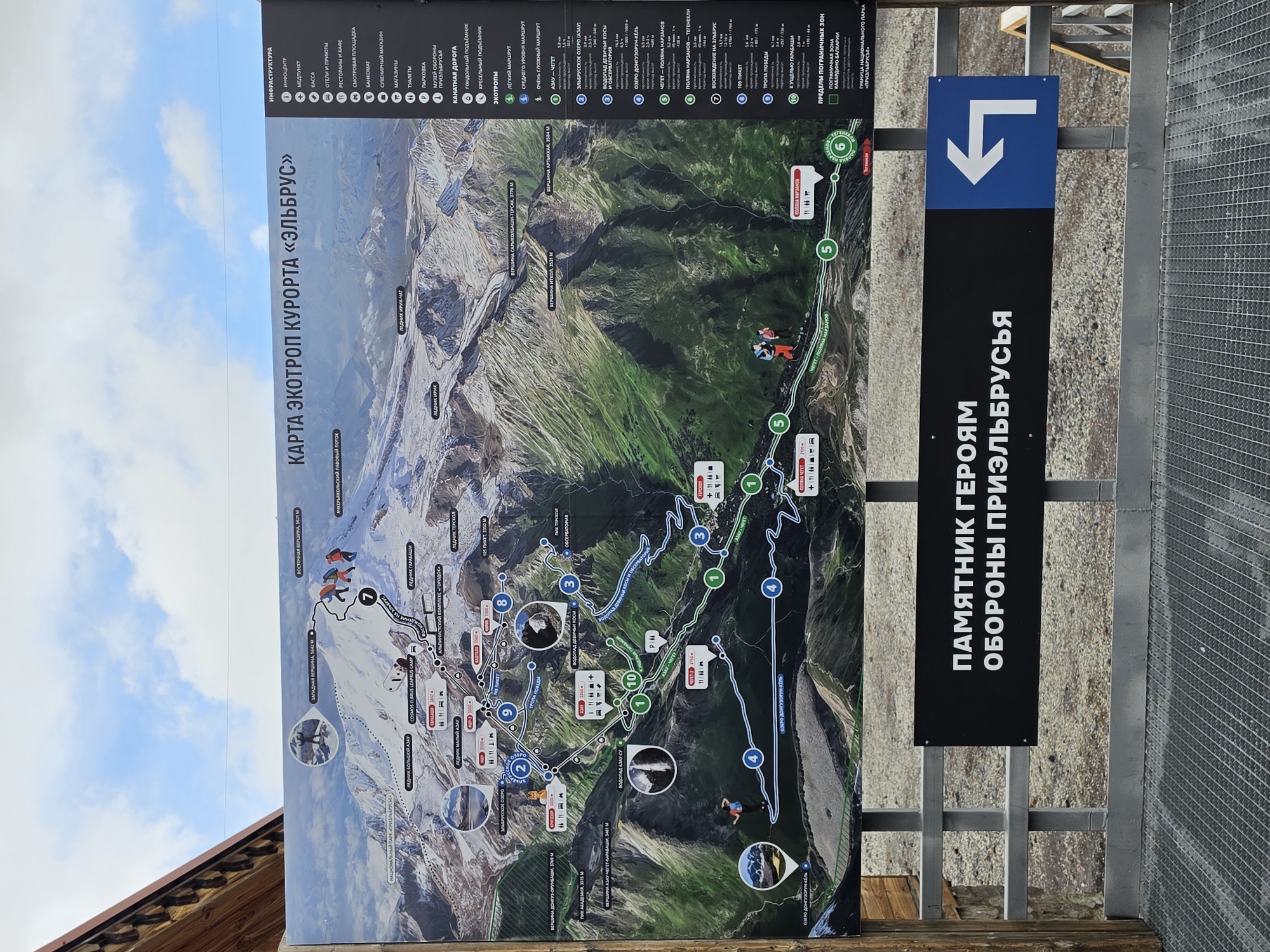 The image shows a large outdoor informational map display at the Elbrus ski resort, located in the village of Terskol, Russia. The map is mounted on a metal frame, with a black sign below it reading "Памятник Героям Обороны Приэльбрусья" (Monument to the Heroes of the Defense of Prielbrusye). A blue and white directional arrow points to the left. The map itself details hiking trails, elevation markers, and points of interest on Mount Elbrus, with a clear sky and mountainous landscape depicted. The scene is set on a cloudy day, with a wooden structure visible on the left and a metal grated walkway in the foreground.