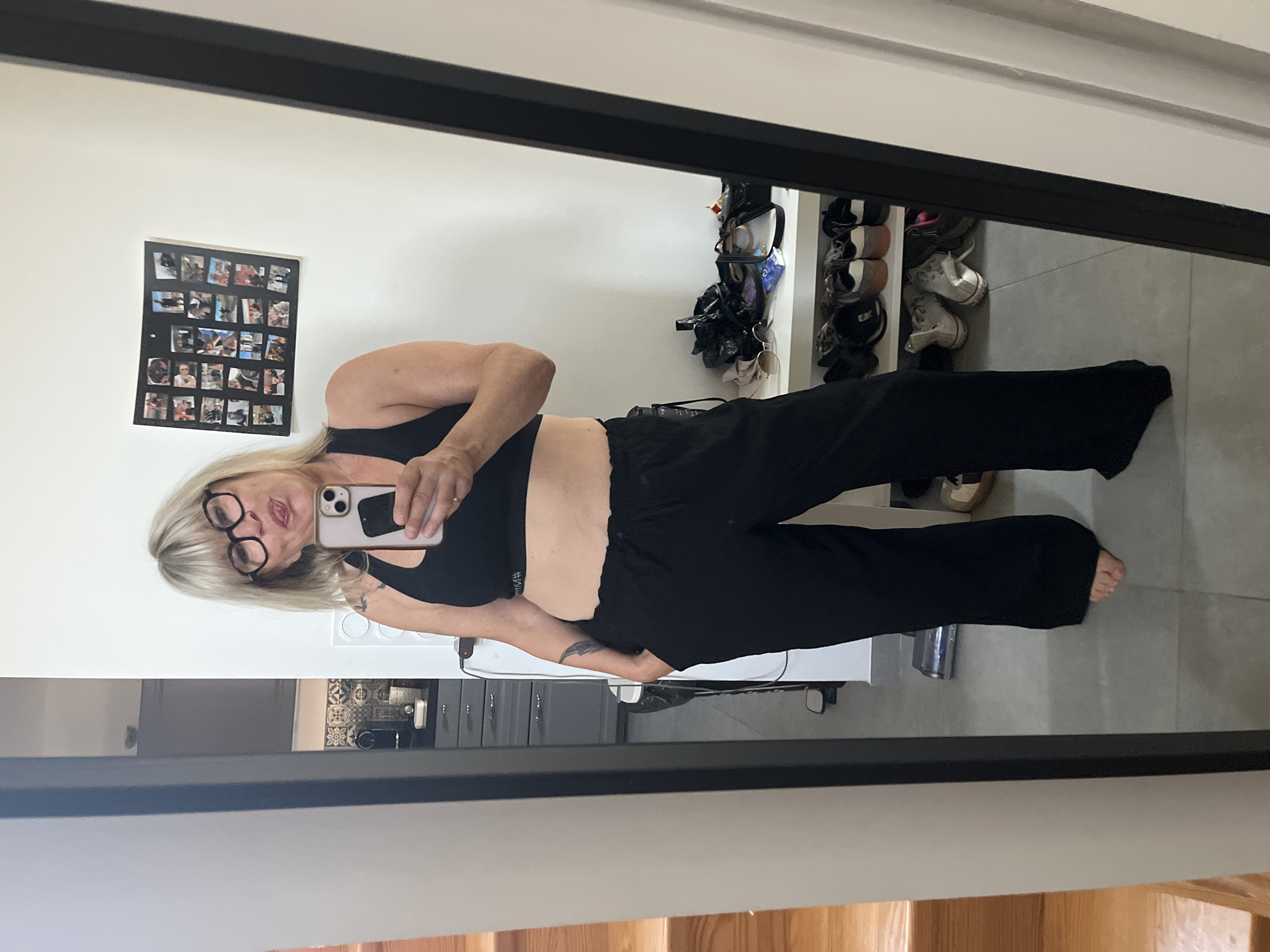 A woman is taking a mirror selfie indoors. She has short blonde hair, is wearing round glasses, a black sports bra, and black pants. She is holding a smartphone in her right hand. In the background, there is a photo collage hanging on the wall, a shoe rack with shoes, a kitchen countertop, and a staircase. The location is not in Chambœuf, France.