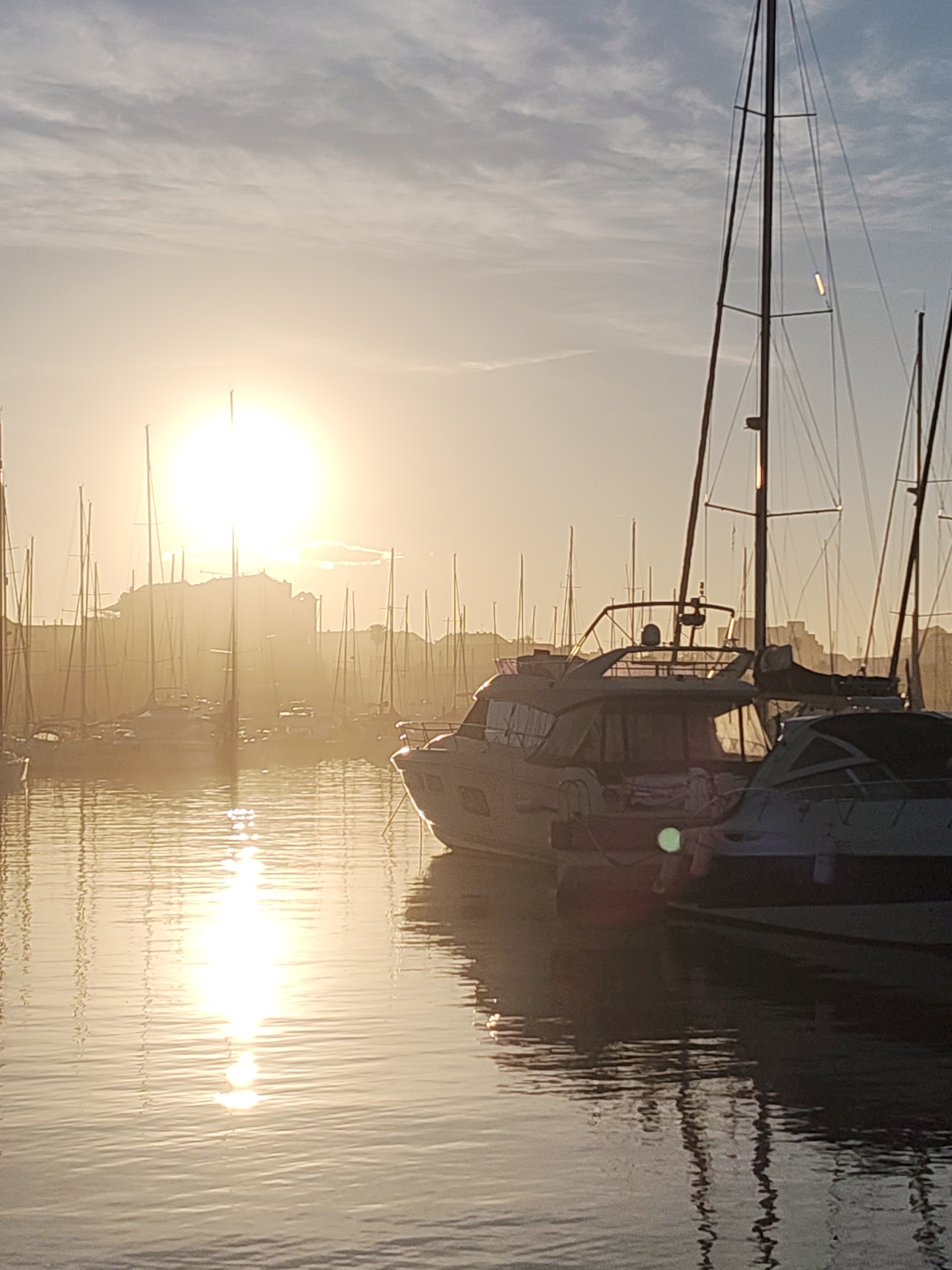 The image depicts a marina at dawn or dusk in Dénia, Spain. Numerous sailboats and motorboats are moored in the harbor. The sun, a bright orb, shines directly into the camera lens, causing a strong glare and silhouetting distant buildings or hills in the background. The water reflects the sunlight, creating a shimmering path across the surface. Two larger motorboats are prominent in the foreground, obscuring the view of other vessels. Tall masts are visible throughout the scene. The overall tone of the image is warm and hazy due to the light.