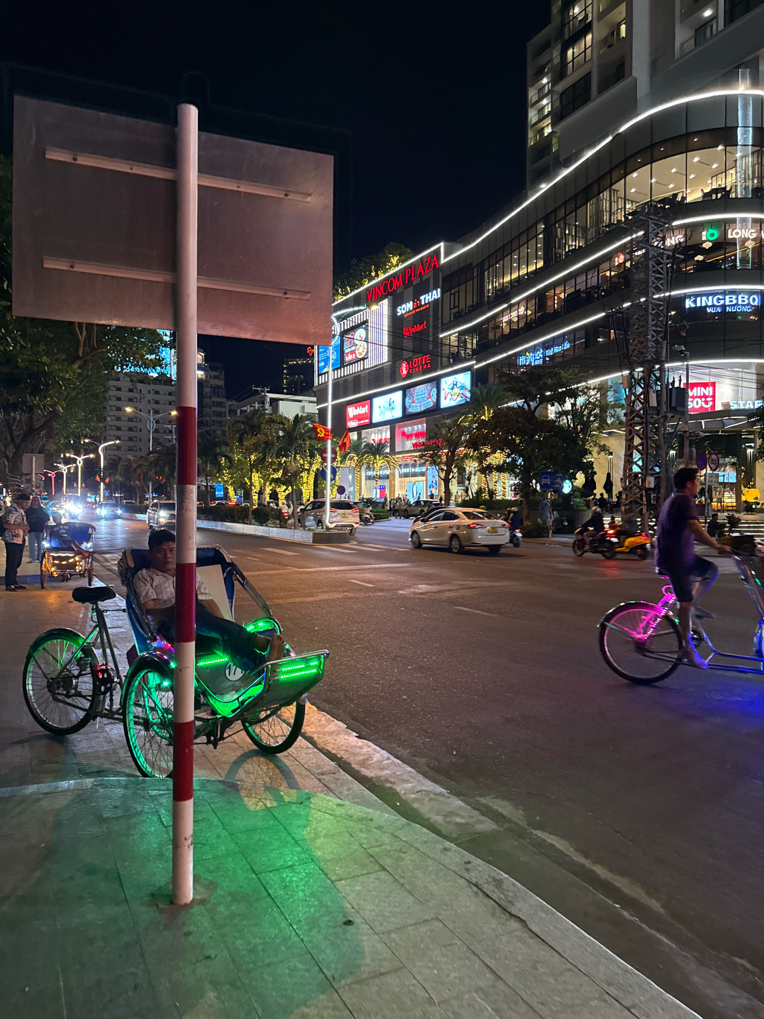 This is a nighttime street scene in Nha Trang, Vietnam, likely in front of Vincom Plaza, a large shopping complex. The foreground features a parked cyclos, a three-wheeled passenger vehicle, illuminated by green LED lights. A man sits in the driver's seat of the cyclos, looking towards the camera. To the left of the cyclos is a bicycle. The street is wet, suggesting recent rain or cleaning, and reflections of lights are visible on the pavement. Cars and motorcycles are present on the street. In the background, the modern architecture of Vincom Plaza is illuminated with bright lights, showcasing various storefronts. The overall atmosphere is urban and lively, characteristic of a city at night.