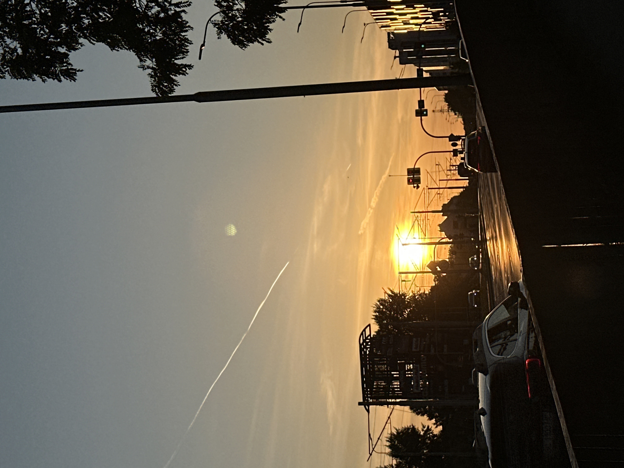 The image captures a street scene in Wrocław, Poland, during sunset. The sun, a bright orb of light, is positioned low in the sky, casting a warm, golden-orange glow across the horizon. A distinct contrail from an airplane stretches across the clear blue sky, arcing from the upper left to the mid-right of the frame.
The foreground is dominated by a dark, silhouetted structure, possibly a railing or barrier, obscuring the lower portion of the image. Beyond this, a street unfolds, lined with traffic lights and street lamps. Several cars are visible on the wet road, their headlights and taillights reflecting the ambient light. One car in the foreground, a white SUV, is partially silhouetted and features prominent roof rails.
In the middle ground, the silhouettes of buildings and trees create a stark contrast against the vibrant sunset. A partially constructed structure, appearing to be a framework of beams, is visible to the left of the frame. To the right, buildings with illuminated facades suggest an urban setting. The overall impression is that of a dynamic city environment transitioning into evening, with the natural beauty of the sunset providing a dramatic backdrop.
