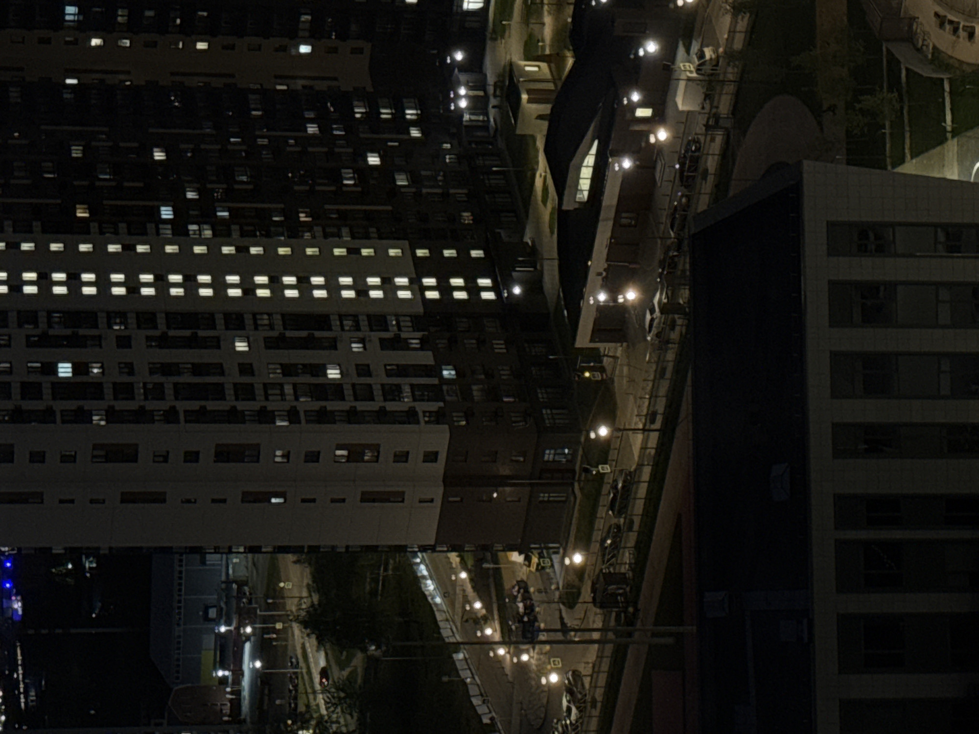 The image shows an aerial view of the city of Lyubertsy, Russia, at night. A large apartment building with many illuminated windows dominates the left side of the frame. Adjacent to the apartment building, a street is lined with streetlights and features moving vehicles. Another tall building is visible to the right, also with illuminated windows. The surrounding environment appears to be a mix of residential and commercial areas, with some green spaces visible. The scene is generally dark, with the lights from buildings and streetlights providing the primary illumination.