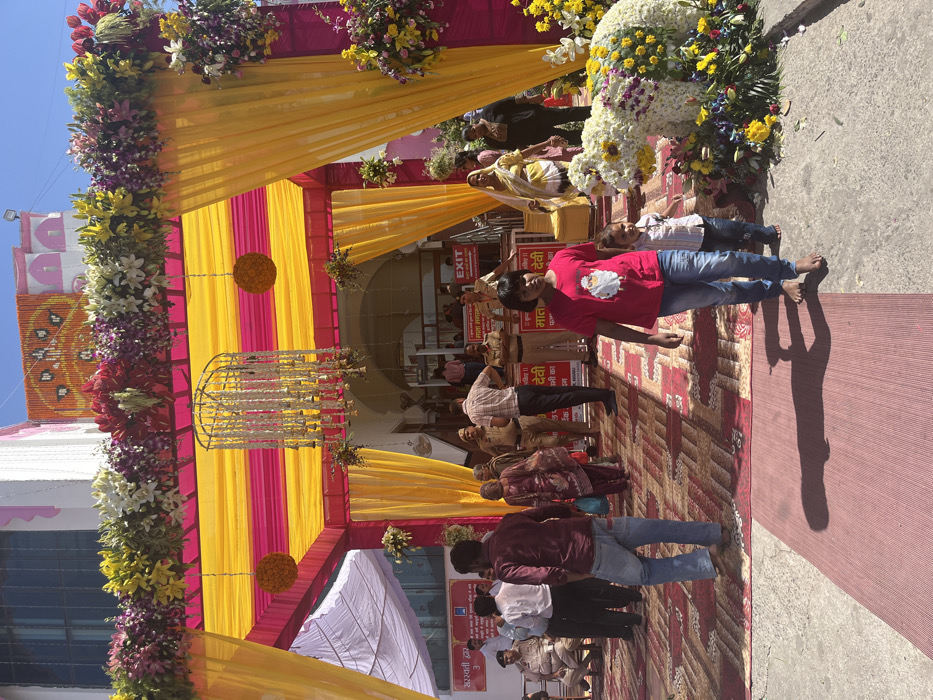 The image shows a scene in Panchkula, India, at a location with coordinates (latitude: 30.724, longitude: 76.86).  The scene depicts a gathering of people in front of a decorated entranceway, likely for a wedding or other celebration. There are several people, mostly men, standing and talking in the foreground, while other people move through the entrance. The entrance is adorned with an elaborate canopy of yellow and pink fabric draped over a metal frame, further embellished with fresh floral arrangements. The entrance is also marked with a red "EXIT" sign. The floor is carpeted with multi-colored patterned rugs.  There are a couple of children, including a young girl in a red shirt and a boy in a blue shirt, interacting with each other. There is also a large flower arrangement in the shape of an animal, likely an elephant, in the right side of the image. The setting is a city building with white walls and a partially visible window. The overall scene is celebratory and festive.