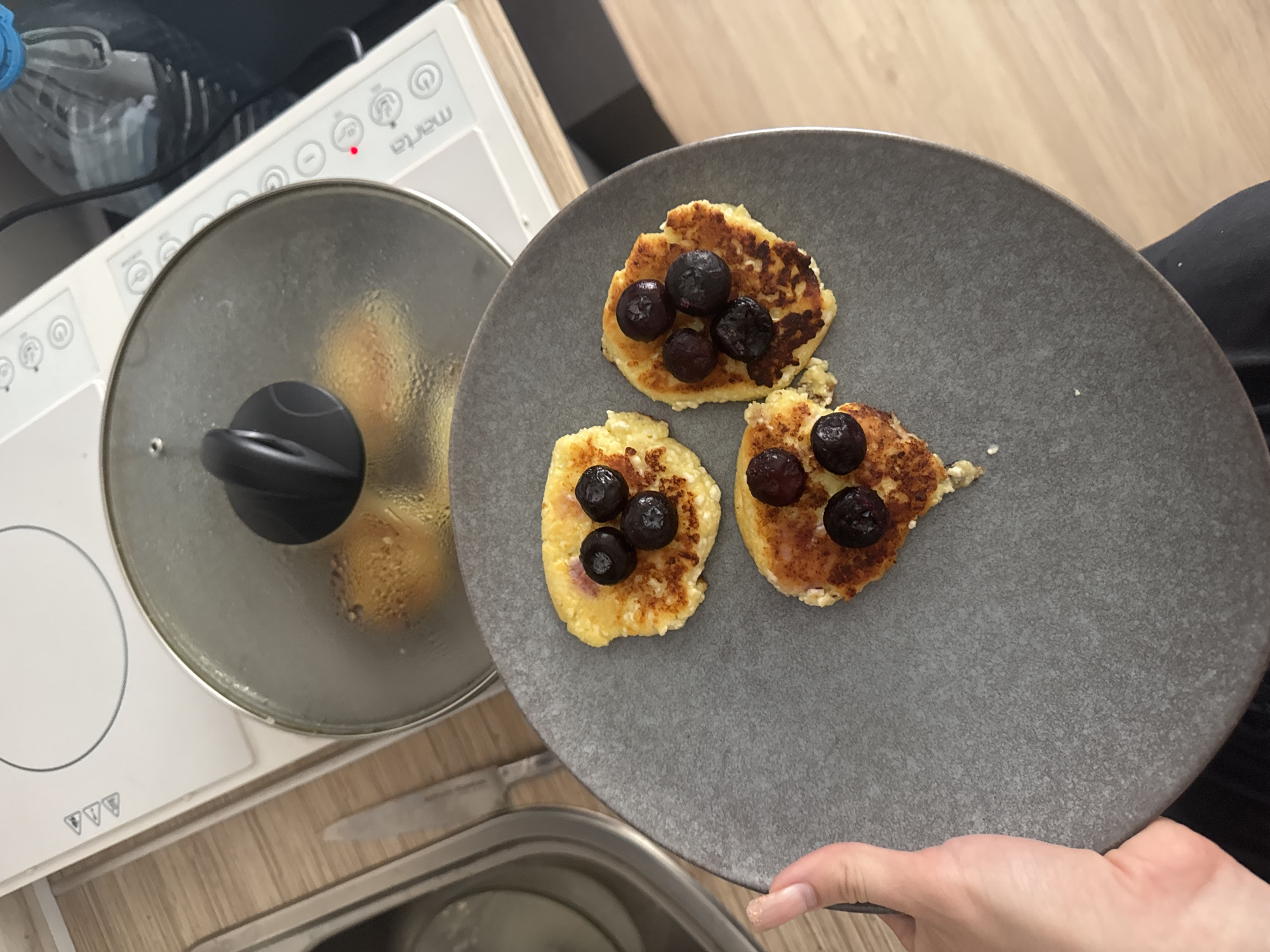 A person's hand is visible on the left side of the image, holding a dark gray plate. On the plate are three small, golden-brown pancakes, each topped with a few dark blue blueberries. In the background, a glass lid covers a cooking surface, with a glimpse of what appears to be more pancakes or fritters underneath. A white electric stove with a glass top is also visible, along with a bottle of water and some kitchen utensils. The scene appears to be in a kitchen setting. There is no information about the location in the city of Perm, Russia.