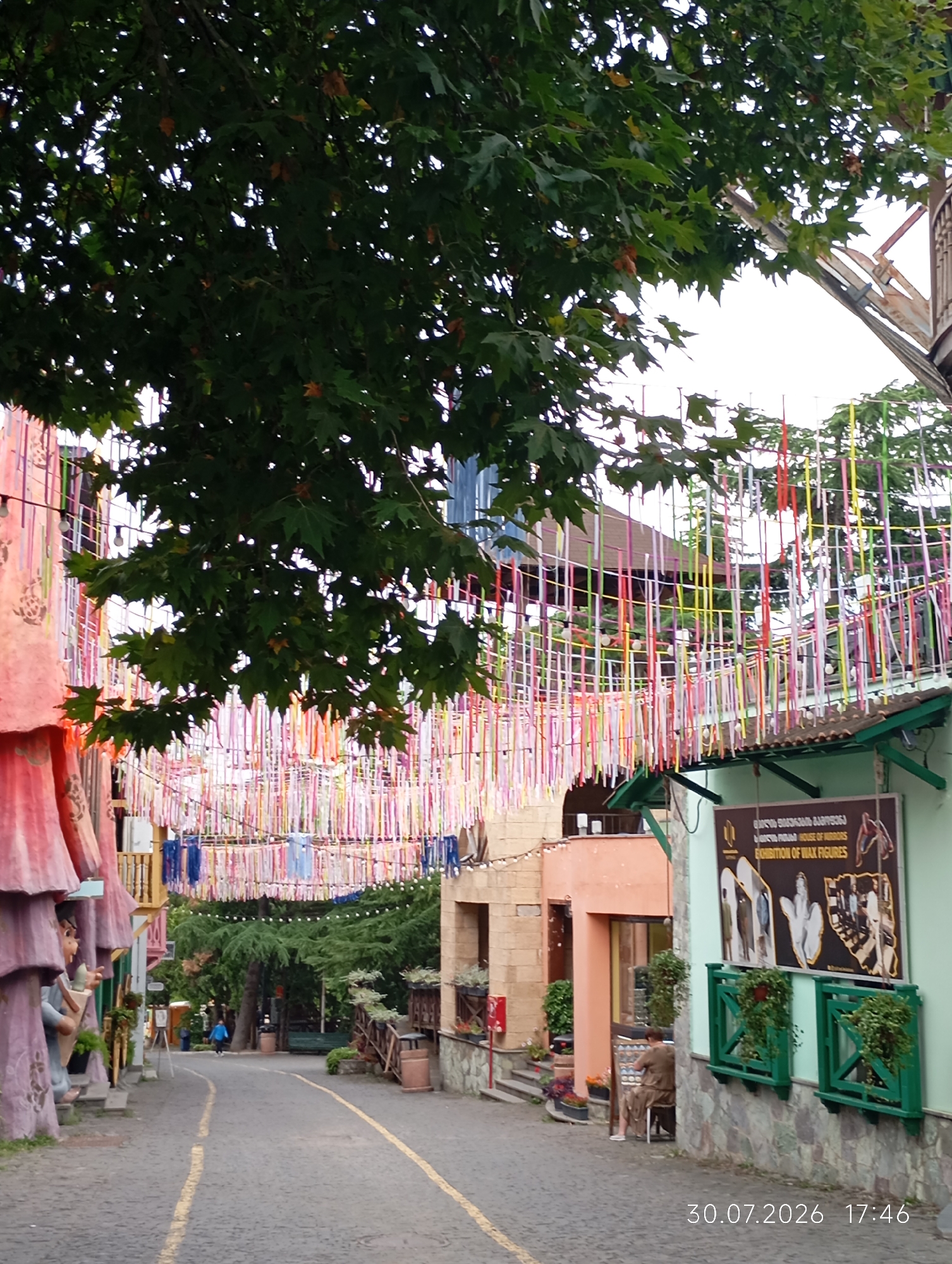 This image captures a street scene in Tbilisi, Georgia, likely on a cloudy day, as indicated by the diffused light. The perspective is looking down a cobblestone street lined with buildings of various architectural styles and vibrant decorations.
On the left, a building features large, colorful, whimsical sculptures that resemble stylized figures or structures. These are pink and purple and appear to be part of the building's facade. Further down the street, more buildings with pastel facades are visible, adorned with a profusion of colorful, thin ribbons strung across the street, creating a festive canopy. Some of these ribbons are pink, red, yellow, blue, and green. String lights are also visible amongst the ribbons.
On the right side of the street, a building is painted light green and has a large, dark sign displayed prominently. The sign features text in Georgian and English, stating "HOUSE OF MIRRORS EXHIBITION OF WAX FIGURES." Below the sign, two green planters with lush green plants are mounted on the wall. The lower portion of this building is clad in rough-hewn stone.
In the background, mature green trees are visible, providing shade and a natural element to the urban setting. There are also glimpses of other structures, including a wooden railing and a bench.
A few people are present in the scene. To the left, a figure is partially visible, appearing to be a statue or decoration on the building's facade. Further down the street, a person in a blue jacket is walking away from the camera. On the right, a person wearing a brown outfit is seated on a stool, facing a small table or easel, appearing to be engaged in an activity.
The street itself is paved with cobblestones and marked with yellow lines. A trash can is visible on the right side of the street. The overall impression is a lively, somewhat eclectic, and colorful street, likely a tourist-oriented area in Tbilisi. The date and time stamp "30.07.2026 17:46" is visible in the lower right corner of the image.