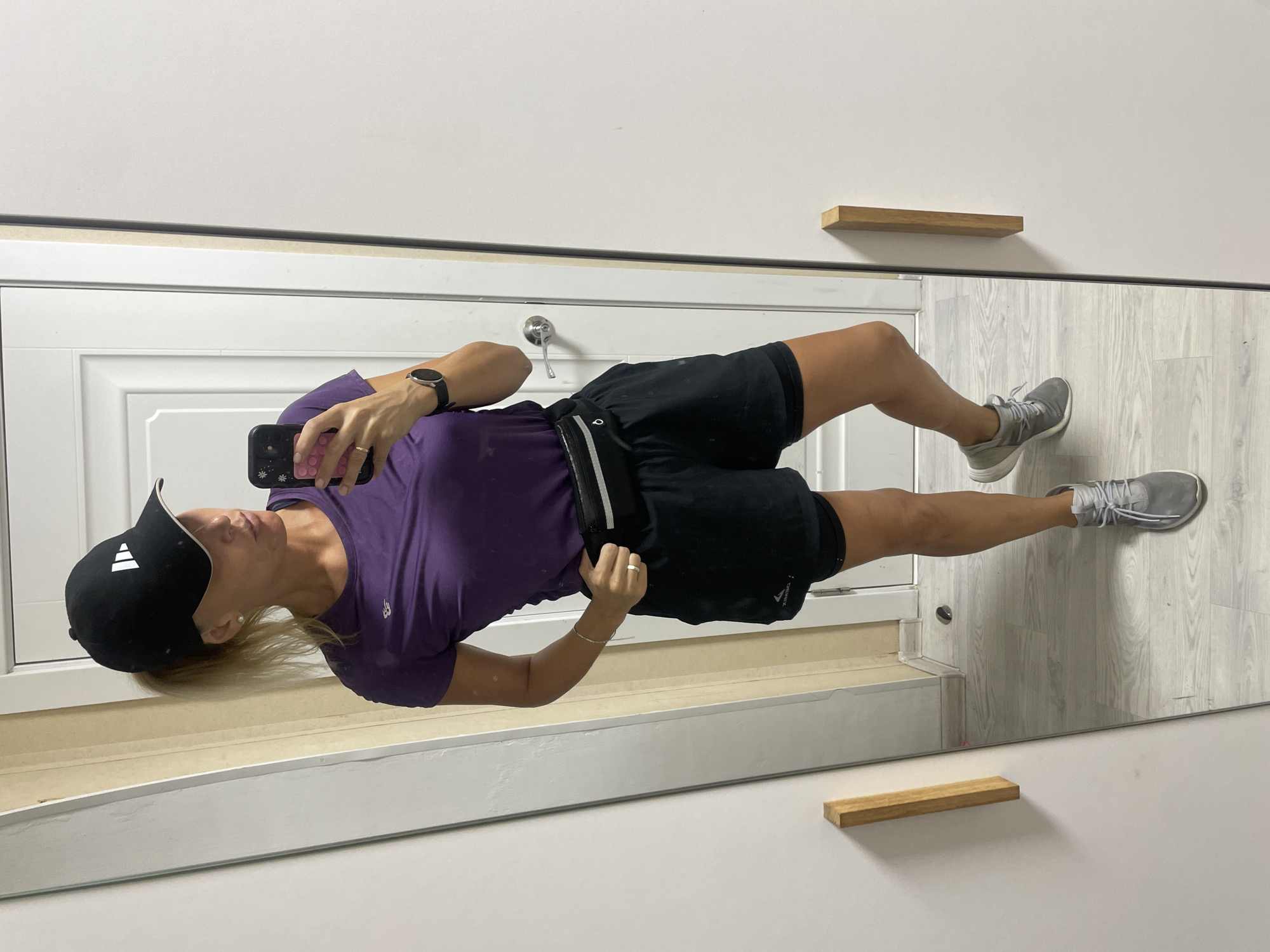 A woman is taking a selfie in a mirror. She is wearing a black Adidas baseball cap, a purple t-shirt, black shorts with a white stripe, and gray sneakers. She is wearing a wristwatch. She is holding a cell phone with a black case. The mirror is framed and there is a white door behind her, as well as two small wooden shelves on the wall. The location is unknown and cannot be identified as Baranovka, Russia.