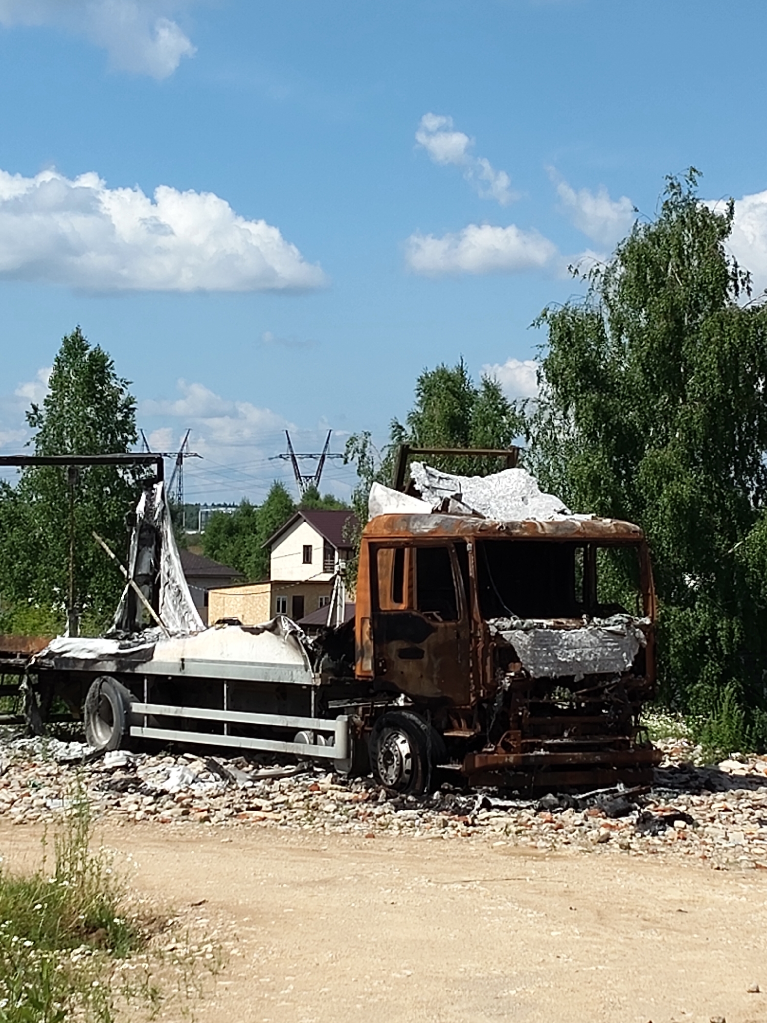 In Chekhov, Russia, a heavily damaged and burned-out truck is situated on a dirt road amidst rubble and debris. The truck's cabin is severely scorched, with melted and warped metal visible. The trailer section appears to have been less affected by the fire but is also damaged, with torn white material draped over it. The surrounding environment is a mix of natural elements and signs of destruction. Green trees and foliage frame the scene, with a bright blue sky dotted with white clouds overhead. In the background, residential buildings and power lines are visible, suggesting the location is near a populated area. The ground is covered in a layer of dirt and broken bricks, indicating past construction or demolition activities. The overall scene depicts the aftermath of a fire or significant incident that has rendered the truck unusable and caused widespread damage to its immediate surroundings.