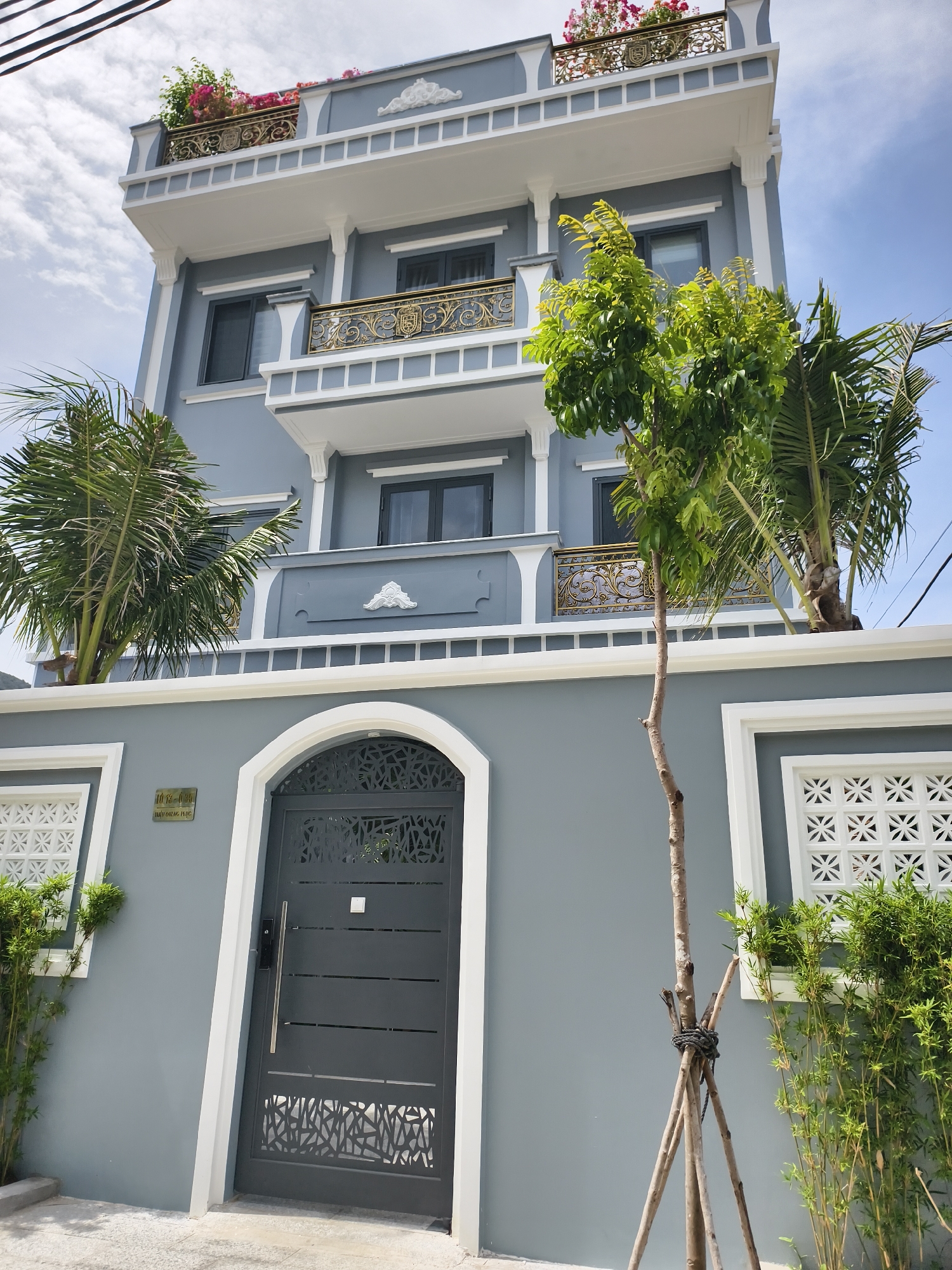 A three-story residential building is pictured on a sunny day in Bac Nha Trang Ward, Vietnam. The building is painted in a muted gray-blue tone, with white trim outlining architectural details. A decorative metal gate, also in a gray-blue hue, stands at the entrance, framed by an arched doorway. Two square windows with white lattice patterns flank the entrance on either side, accompanied by shrubbery. Two palm trees and a slender tree, supported by stakes tied with rope, are positioned in front of the building. Balconies with ornate gold railings are visible on the upper floors, with the top level featuring a rooftop garden with pink flowers. A street address plaque is mounted on the wall near the entrance. Overhead, electrical wires stretch across the upper portion of the image against a partly cloudy blue sky. The pavement in front of the building is visible.