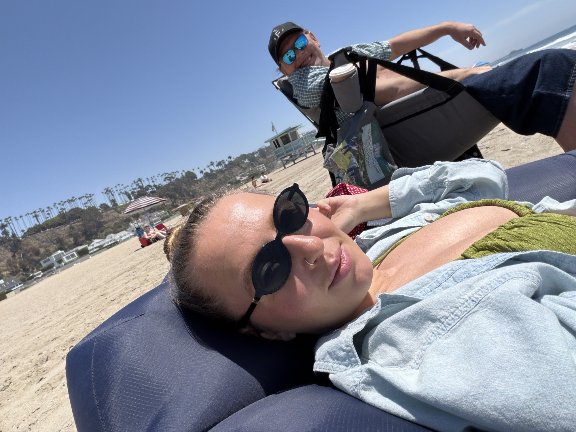 The image shows two people on a beach in Santa Monica, California. In the foreground, a woman is lying on a blue inflatable lounger, wearing a green top, a light blue denim shirt, and round sunglasses. She has a nose ring and her hair is pulled back. Behind her, a man is sitting in a gray folding chair. He wears a plaid shirt, dark blue shorts, a black cap with white lettering, and sunglasses with blue lenses. A cup in a holder and a tote bag are attached to his chair.
The background includes a sandy beach, a lifeguard tower, the ocean, and a hillside with palm trees and buildings. Some other people are visible on the beach. The sky is clear and blue.
