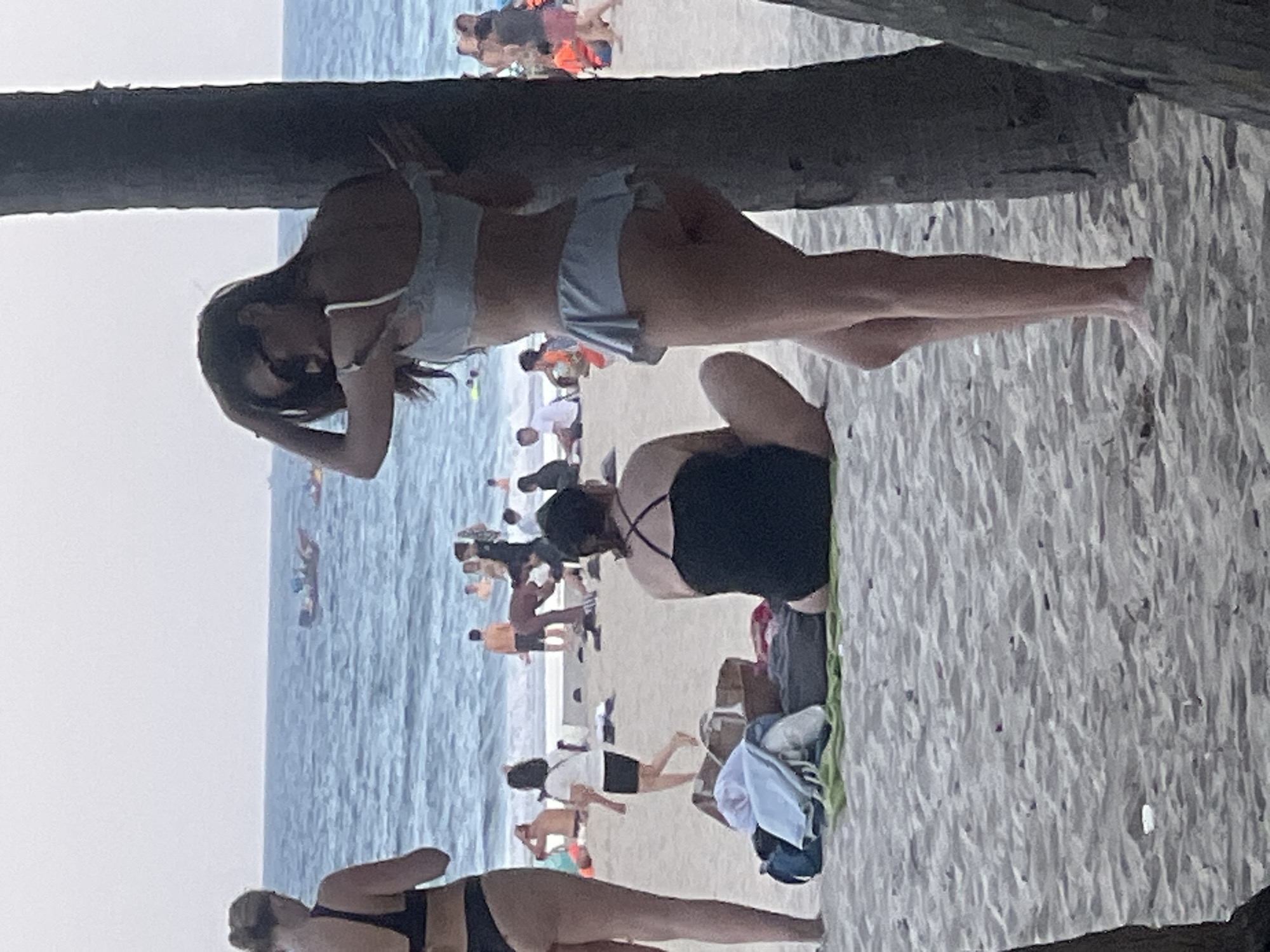 The image depicts a beach scene in Da Nang, Vietnam, with several individuals present. In the foreground, a woman in a light-colored bikini is leaning against a tree trunk, looking towards the sea. To her left, another woman in a black bikini stands facing away from the camera. Further back, a woman in a black one-piece swimsuit is seated on the sand, facing towards the ocean. Several other beachgoers are visible in the mid-ground, some walking and others sitting. The sea stretches out in the background, with a few boats on the horizon. The beach itself is sandy, with scattered beach towels and bags. The sky is overcast.
