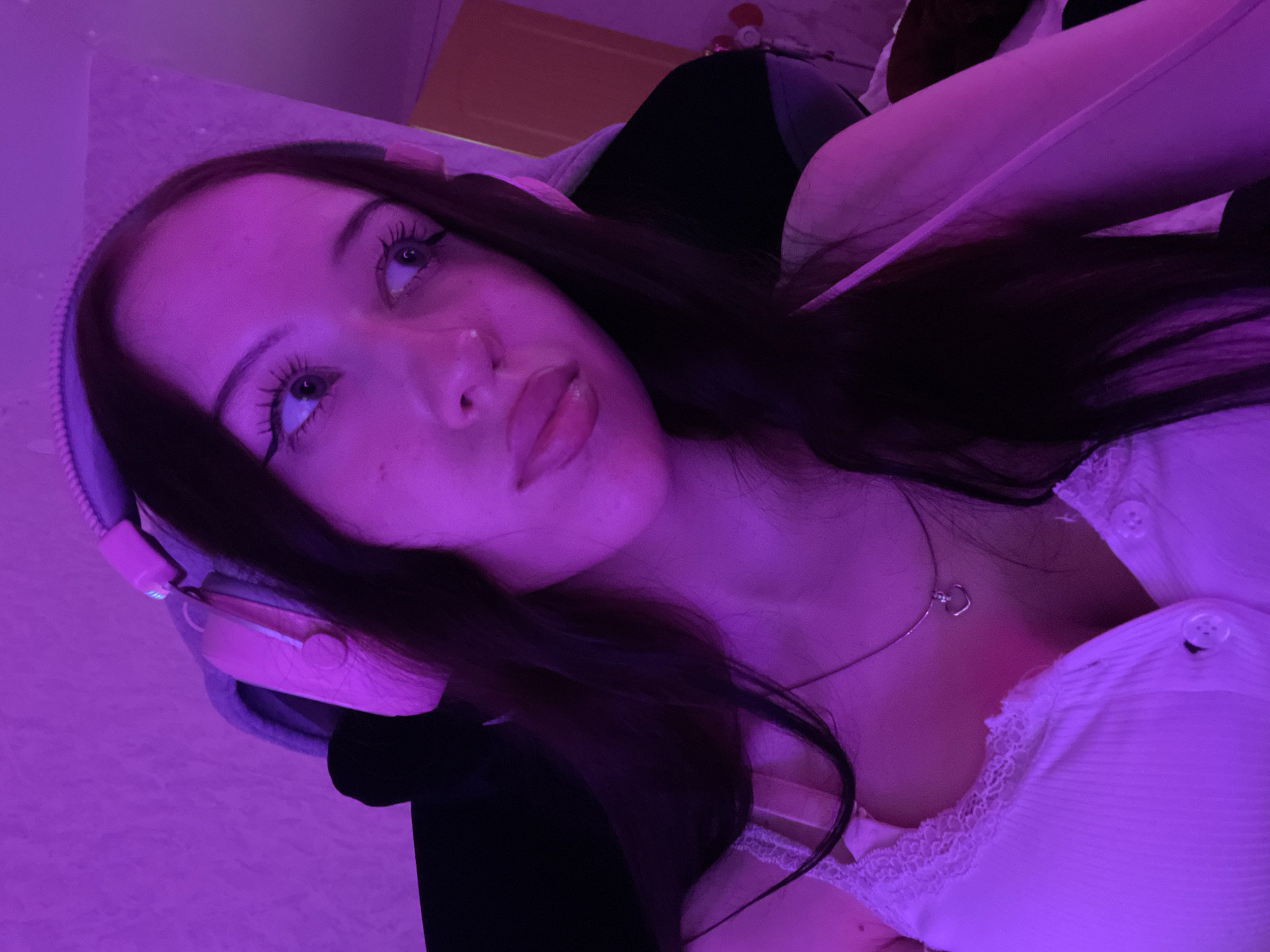 A young woman with long, dark hair is wearing pink headphones and a white tank top. She has on eye makeup and a necklace with a heart-shaped pendant. She is looking off to the side with her mouth slightly open. The scene is lit with purple light, creating a moody atmosphere. The background appears to be a room with a wall and possibly some furniture. There is no specific information in the image to place it in Votkinsk, Russia.