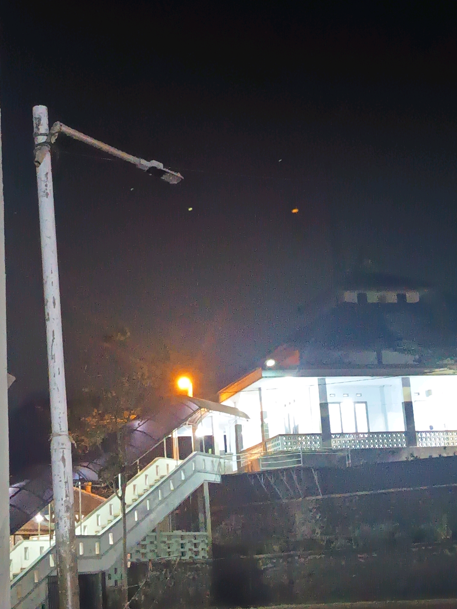 This is a nighttime, outdoor, medium-shot photograph taken from a low angle. The image captures a building with a white facade, illuminated by interior lights, and a prominent outdoor staircase leading up to it. The staircase is made of white concrete and features railing on both sides. The building appears to be a public structure, possibly a mosque or community center, given the architectural style and the presence of what looks like a minaret or tower-like structure on the right side, partially obscured by shadows. A streetlamp, emitting a warm orange light, is visible on the left side of the staircase. The background is dark, indicating a nighttime scene, with a few distant lights or objects appearing as small, bright specks in the sky. The overall scene suggests a quiet urban or semi-urban environment in Sumedang, Indonesia, likely taken during evening hours when the building is lit for use.