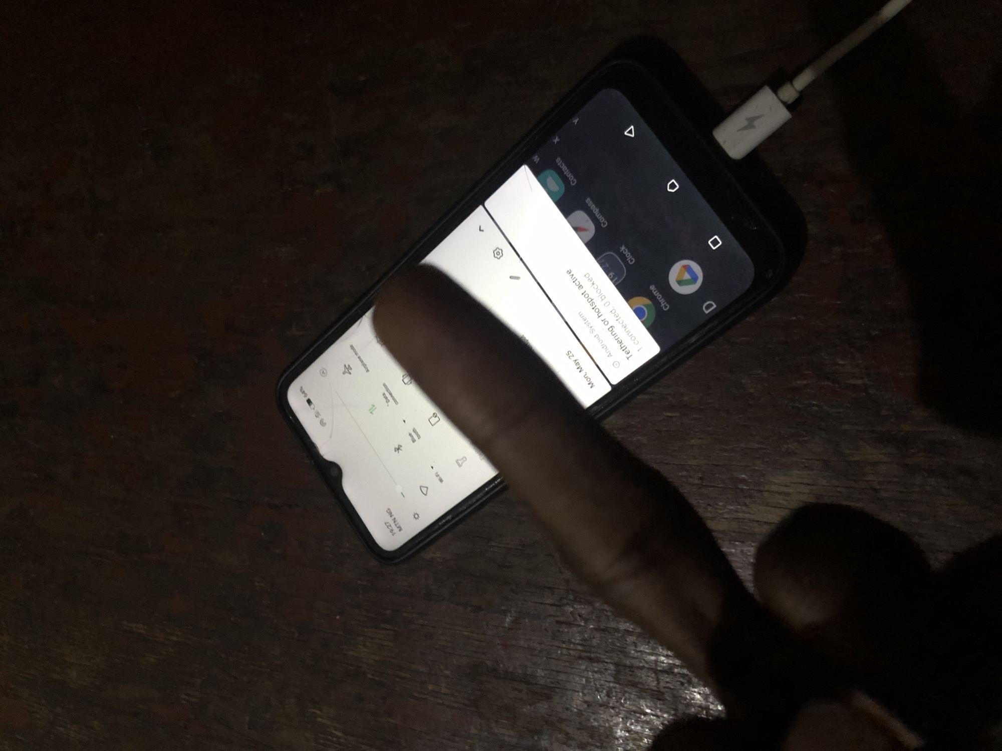 The scene presents a close-up view of a smartphone placed on a dark wooden surface. A charging cable is plugged into the phone. On the screen of the phone, a notification is visible stating "Tethering or hotspot active, 1 connected, 0 blocked" and below it, "Mon, May 25." A finger is hovering over the screen, presumably interacting with the interface. The smartphone displays app icons for Chrome, Clock, Compass, and Contacts. The device's status bar indicates a time of "19:27" and a network named "MTN NG." Airplane mode and data connection settings are also visible on the screen. The surrounding environment is dimly lit, focusing attention on the phone and the user's interaction with it. The location is Unknown, Unknown.