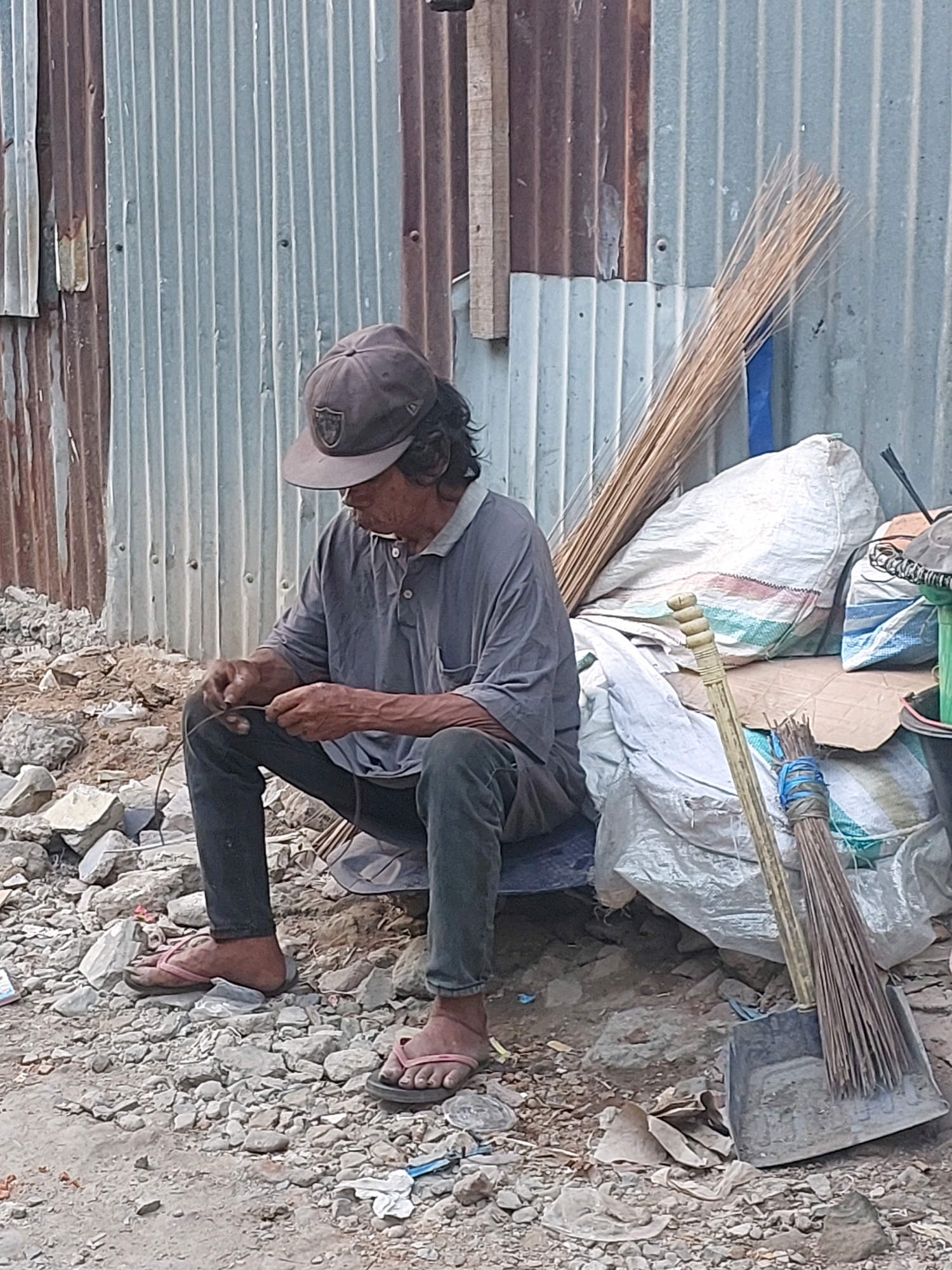 A man sits on rubble in Ambon, Indonesia, facing a corrugated metal wall. He wears a gray polo shirt, dark pants, a baseball cap, and flip-flops. His hands are occupied with a thin wire. Beside him are large sacks, a broom, and a dustpan. The ground is covered in debris and rocks.