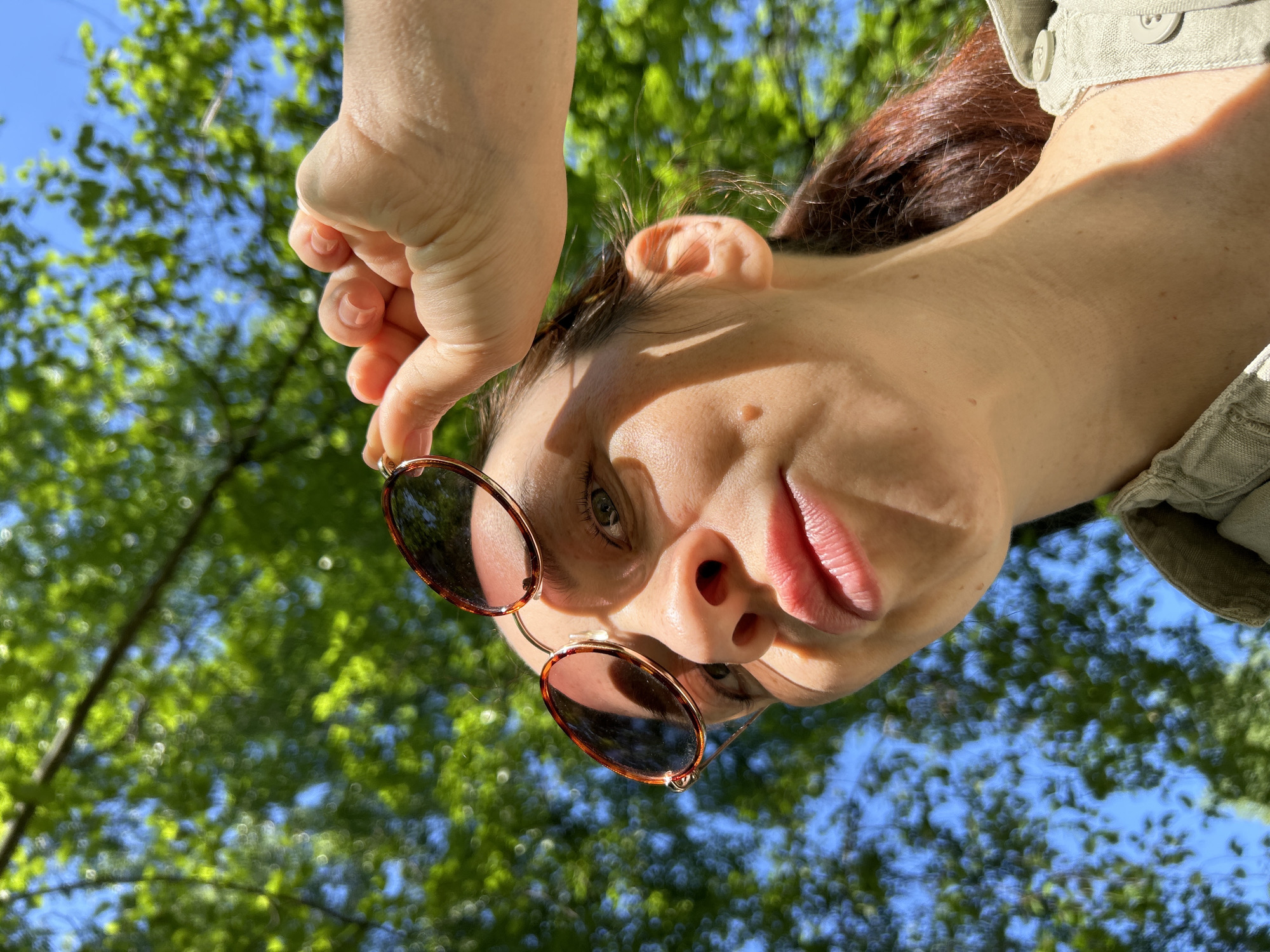 A low-angle shot depicts a woman holding sunglasses, with trees and a blue sky in the background. Her head is tilted downwards, and she is wearing a light-colored collared shirt. The location is implied to be Chaikovsky, Russia, though there are no discernible markers in the image to confirm this detail.