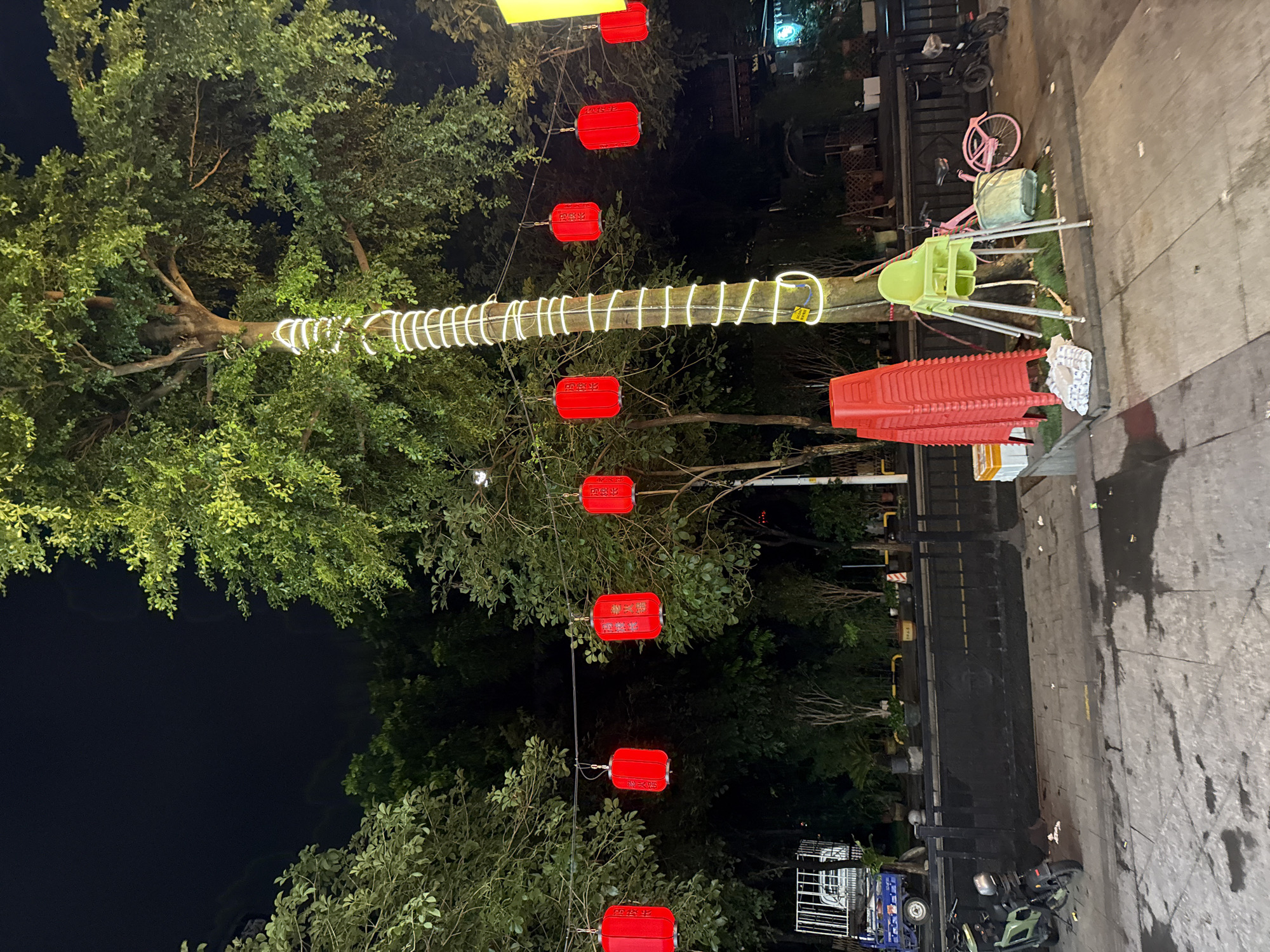 This image captures a nighttime scene in Haizhu District, China, likely in an outdoor public space like a street or park. The primary subjects are a large, leafy tree adorned with a string of white, coiled lights and several red, rectangular lanterns hanging from wires. The lanterns appear to be decorative, possibly for a festival or event. In the background, a street or sidewalk is visible, with some debris scattered on the wet ground, suggesting recent rain or a damp environment.
Several objects are present: a stack of red plastic chairs, a light green child's high chair, and a pink bicycle with a basket. Further in the background, there are more decorative elements, potentially part of a market or seating area, and a small vehicle. The lighting suggests it is nighttime, with artificial lights illuminating the scene. The overall mood is festive due to the lanterns, but the scattered debris and wet ground hint at a more mundane, possibly transient, setting. No people are clearly visible in the image, focusing the attention on the decorations and the environment.