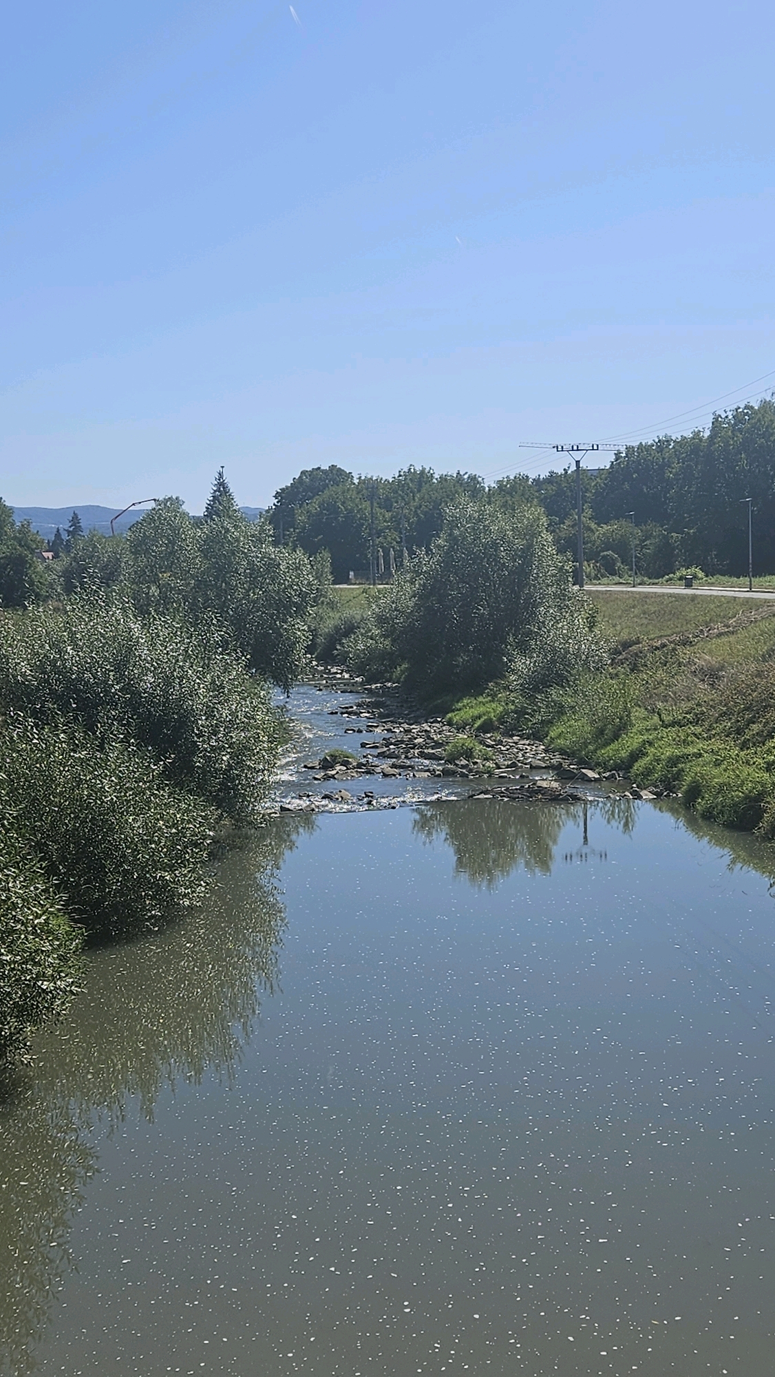 The image depicts a tranquil natural scene of a river flowing through a lush green environment under a clear blue sky. The river, shallow and clear, meanders through a rocky bed, with its banks densely populated by various trees and bushes. The water surface reflects the surrounding greenery and the sky, creating a serene and picturesque view. In the distance, a few structures and power lines are visible, hinting at human presence on the periphery of this natural landscape. The image appears to have been taken on a bright, sunny day. The exact location in Prešov, Slovakia is not identifiable from the image alone.
