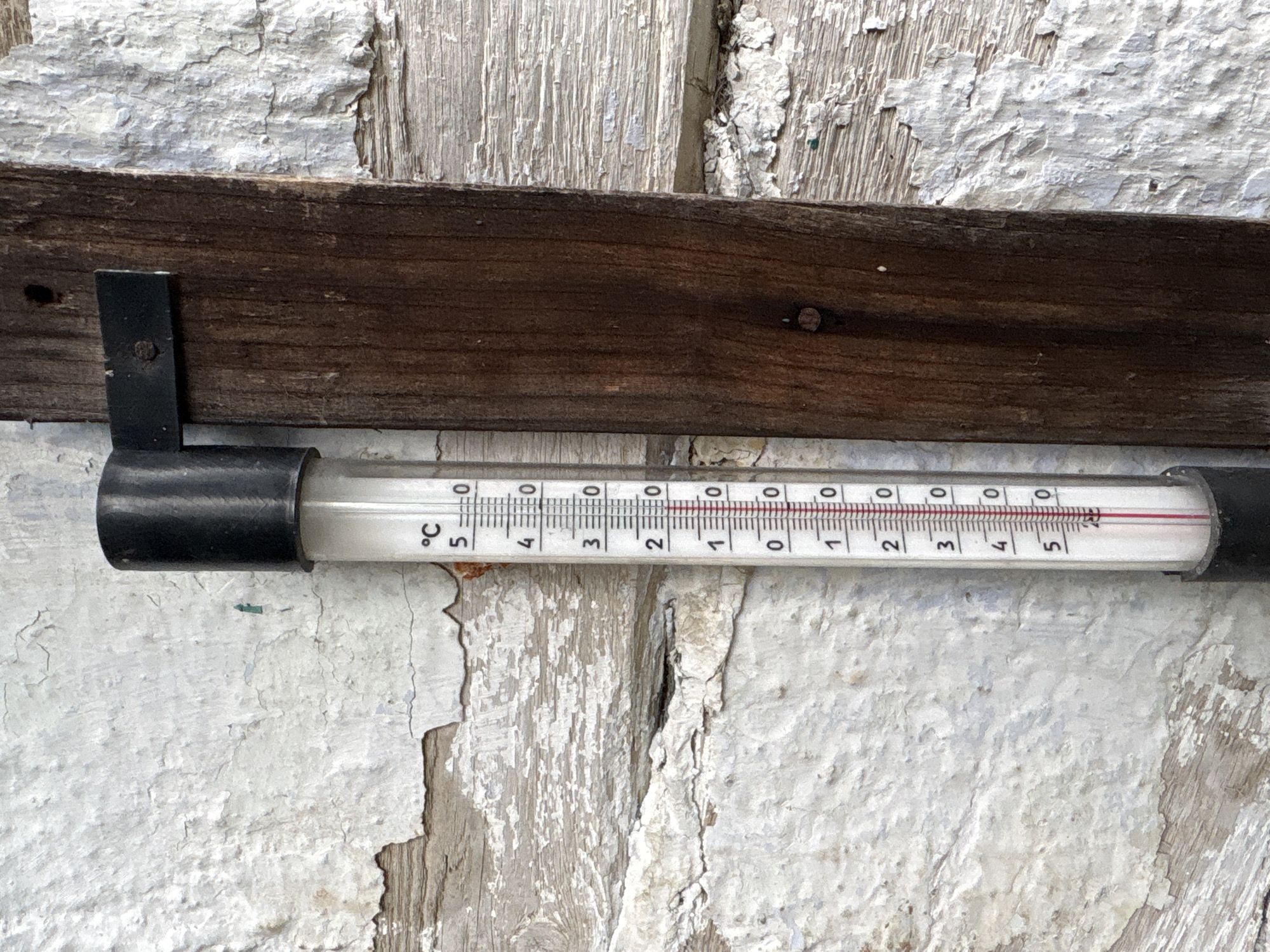The image shows a thermometer attached to a weathered wooden post. The thermometer displays a temperature of approximately 5.7 degrees Celsius. The background consists of a textured, peeling white wall and a rustic wooden plank. The scene appears to be outdoors, possibly in a rural setting or an older part of a town. No people or specific landmarks are visible. The thermometer is the primary subject, indicating a cool ambient temperature.