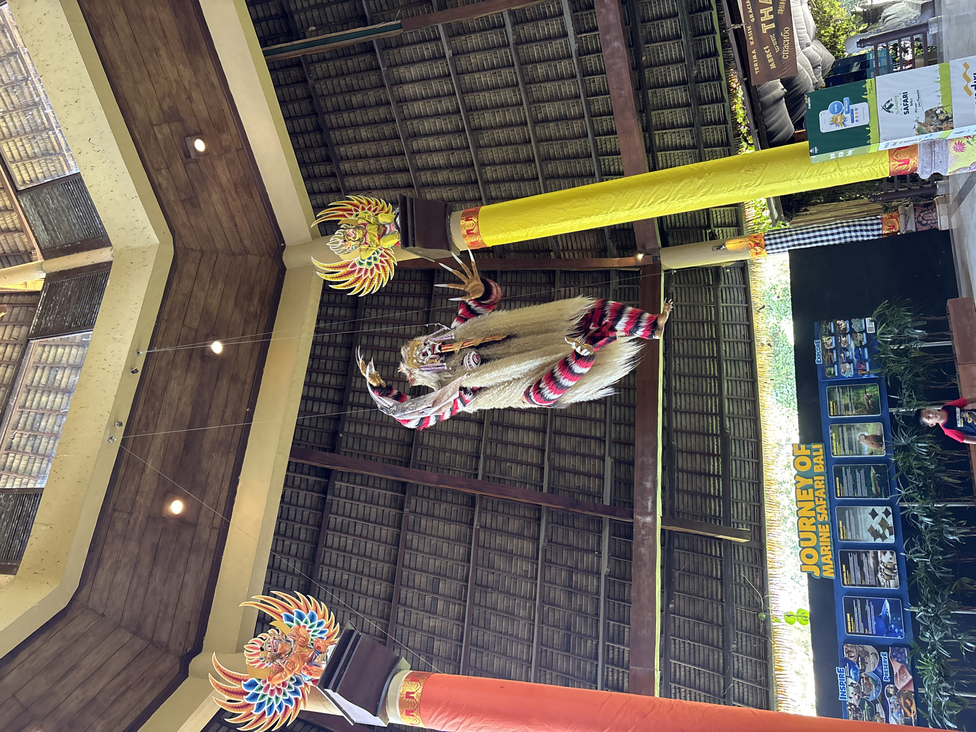 This photograph captures the interior of a large, open-air building, possibly a lobby or entrance area, in Blahbatuh, Indonesia. The architecture features a high, thatched roof supported by wooden beams and pillars. Dominating the scene are two large, decorative statues hanging from the ceiling: one on the left depicts a creature with colorful wings, possibly Garuda, and another, larger figure in the center appears to be a demon or mythical being, adorned with white fur, red and white striped garments, and fierce facial features. In the background, a sign reads "JOURNEY OF MARINE SAFARI BALI," indicating the location is likely within the Bali Safari and Marine Park. Below the sign, informational displays about different "zones" such as "Ocean Zone" and "River Monster" are visible. A young boy in a red shirt is standing near the bottom center of the frame, looking towards the camera. Several potted plants are arranged in front of the informational displays. The overall impression is one of a vibrant, culturally rich environment, characteristic of tourist attractions in Bali.