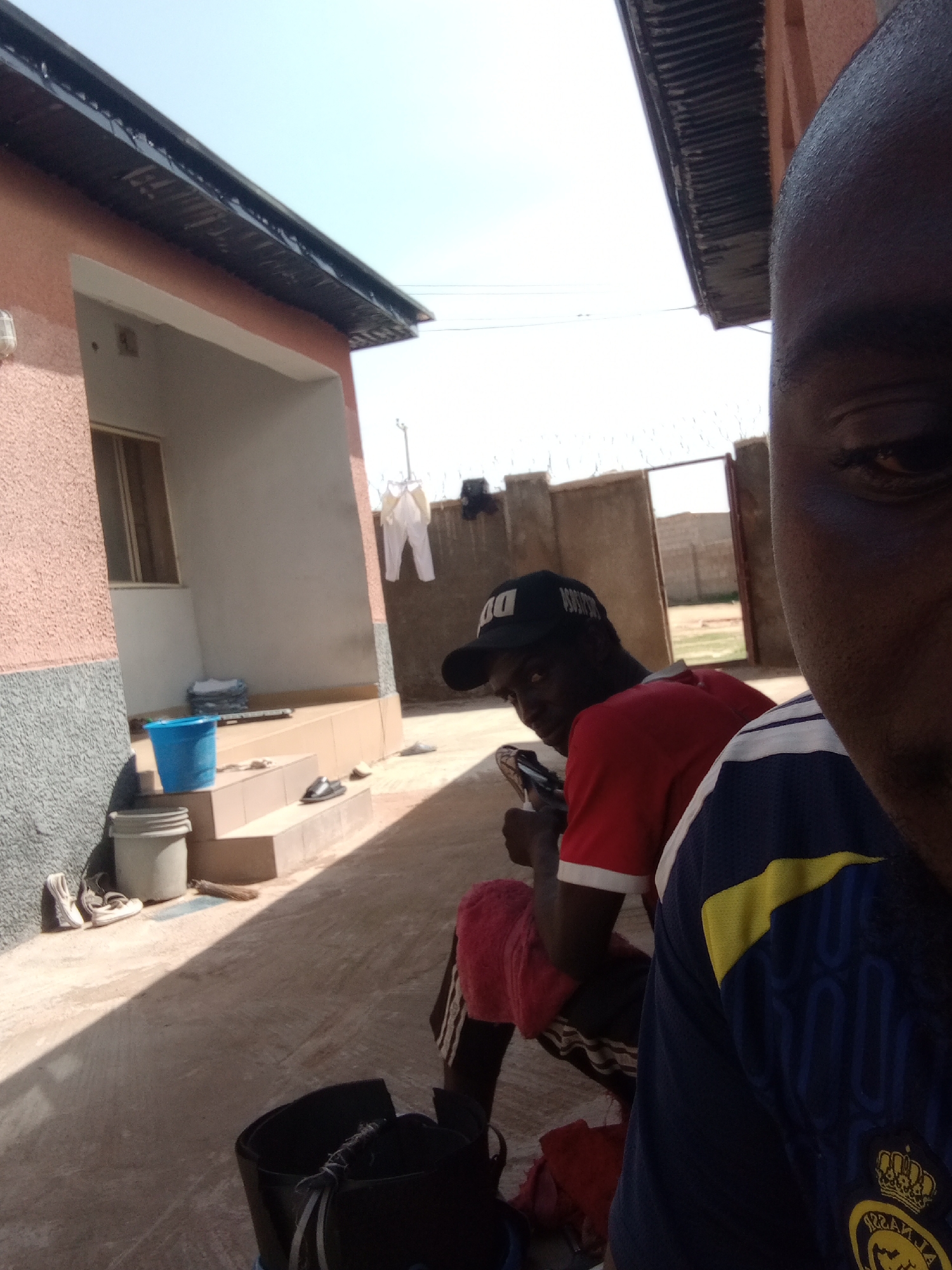 The image depicts an outdoor scene likely in a residential area. In the foreground, the face of a person is partially visible. In the middle ground, another person is seated, wearing a red t-shirt and a black baseball cap. They appear to be holding a phone or similar object. Behind them, a doorway leads to an open area, possibly a yard or street, with a fence in the background. The environment includes a building with an overhang and a window, as well as steps leading up to an entrance. Various objects are scattered around, including buckets, shoes, and laundry hanging on a line. The lighting suggests it is daytime, with sunlight casting shadows on the ground.