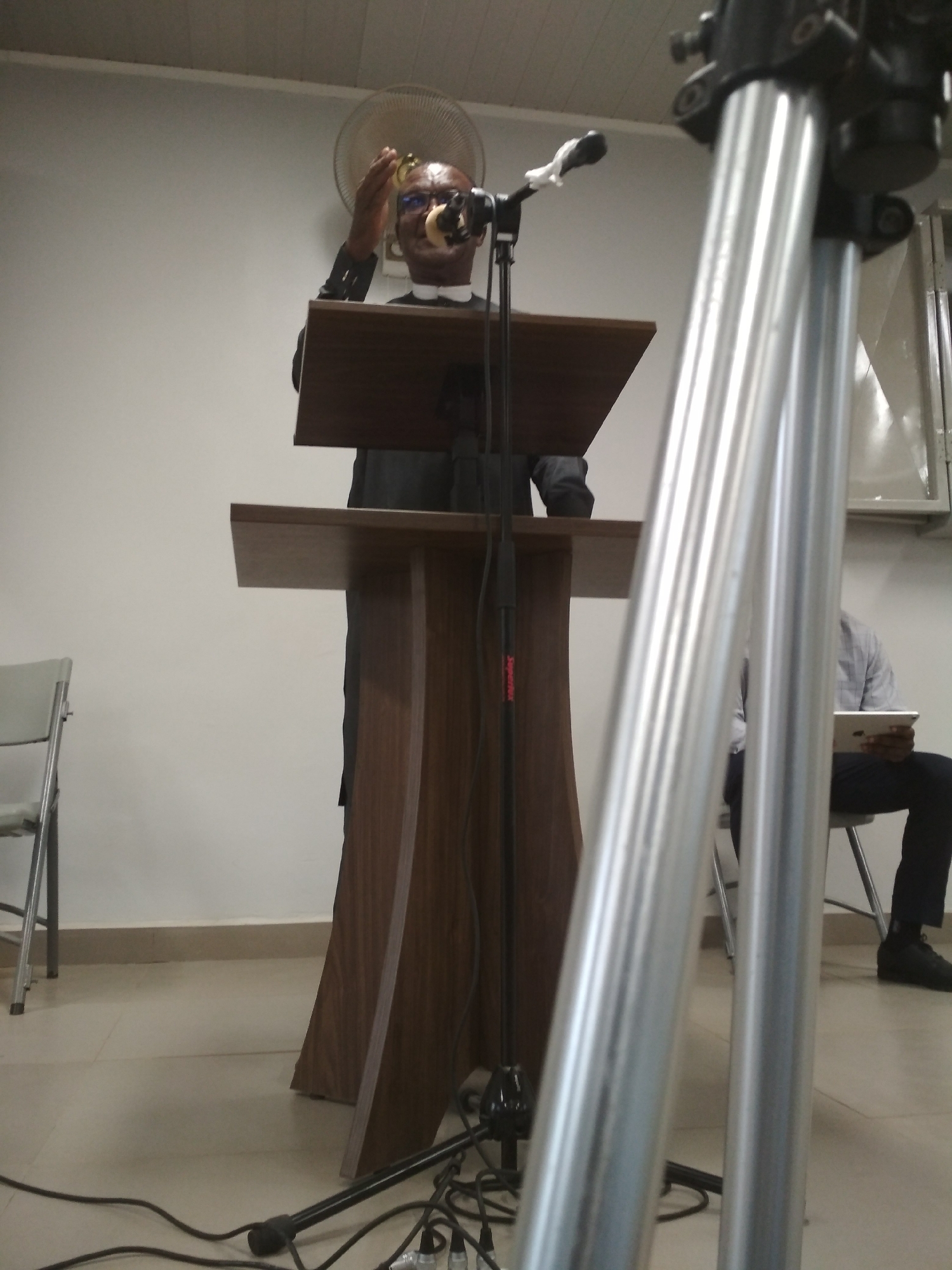 A man in a black suit stands at a wooden podium in what appears to be an indoor venue. He is wearing glasses and gesturing with his right hand while speaking into a microphone. Behind him is a white wall with a fan mounted on it. To the right, partially obscured by a silver tripod, is a person sitting and looking at a tablet. The floor is tiled and there are wires snaking along the floor near the podium. The location is Gura Top, Nigeria.