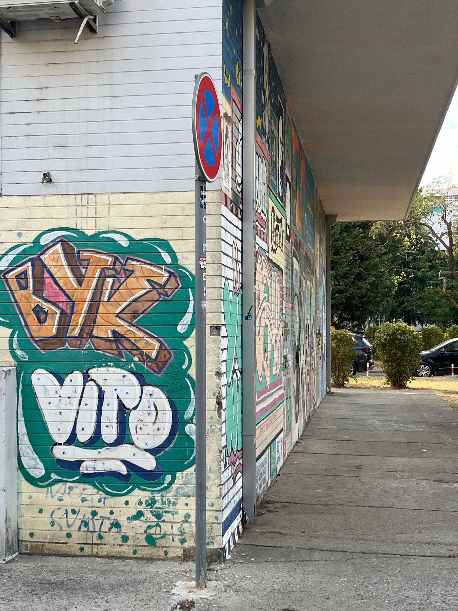 A wide-angle, ground-level outdoor shot shows a building with a prominent graffiti-covered wall. The graffiti on the left is in vibrant green and orange, with blocky lettering that reads "BYZ" and "VIR." The wall is made of light-colored brick. To the right of the graffiti, a cylindrical pole supporting a "no parking" sign is visible. Further to the right, the building's wall is covered in a colorful, multi-panel mural depicting various images and text in a comic-book style. The mural extends down the length of the building, which features a projecting concrete overhang. A concrete pathway runs along the base of the building, leading into the distance. Behind the pathway and building, lush green trees and bushes form a backdrop. Two dark-colored cars are parked in the background, partially obscured by foliage. The scene appears to be a street or alleyway in a city, possibly Podgorica, Montenegro, given the context. The lighting suggests it is daytime, with sunlight casting shadows.