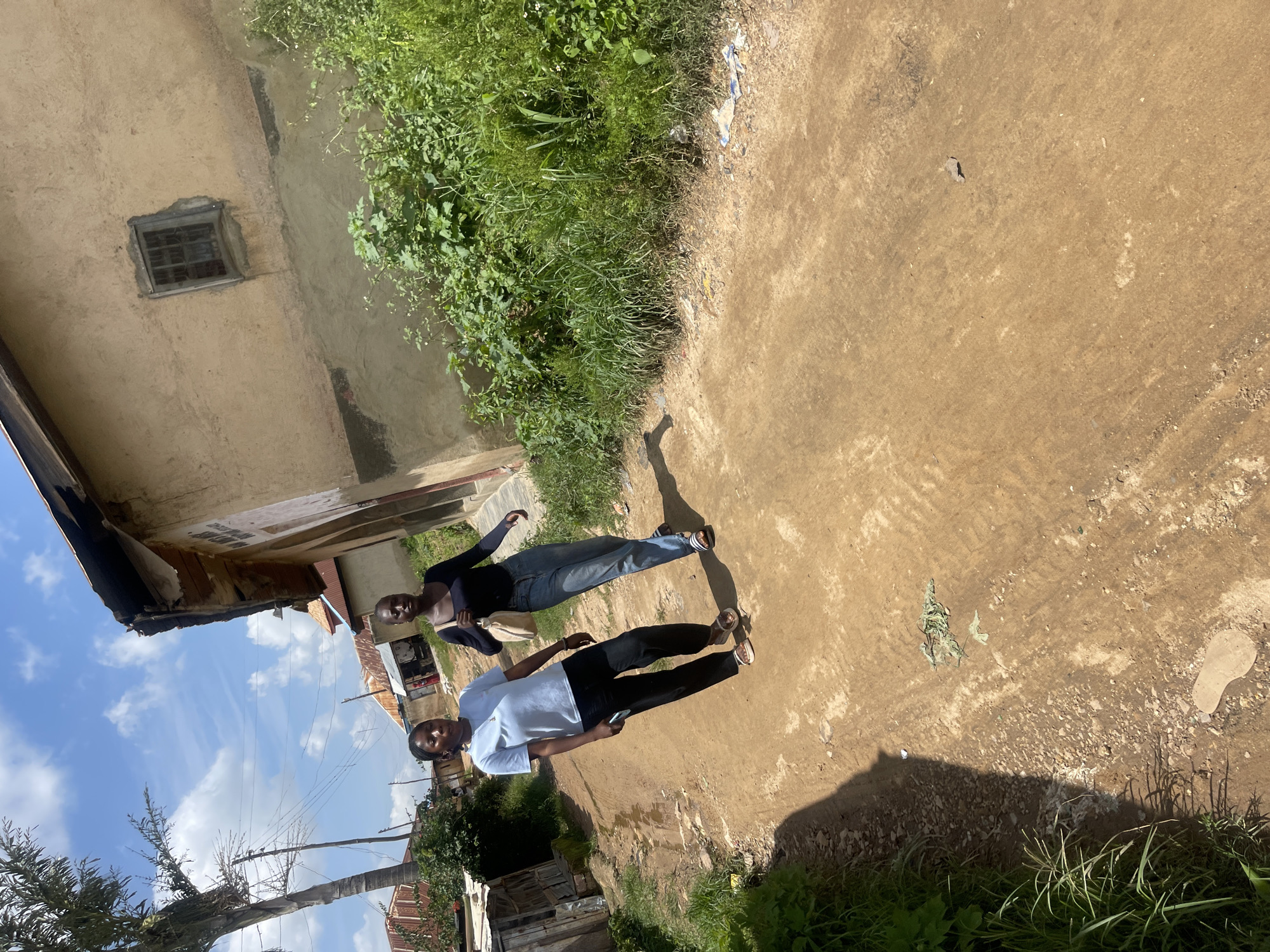 This media file shows two young women standing on a dirt path in Rafin Par, Nigeria. One woman is wearing a black long-sleeved shirt and jeans, while the other is wearing a light blue t-shirt and black pants. They are both looking at the camera with neutral expressions. The scene appears to be outdoors, with a building on the left and lush green vegetation on the right. The sky is visible in the background, with some clouds and power lines. The ground is a dusty, reddish-brown dirt path. The sunlight suggests it is daytime, likely late morning or early afternoon, given the shadows cast by the people. There is no visible text in the image. The overall impression is a casual snapshot of two individuals in a simple, rural setting.