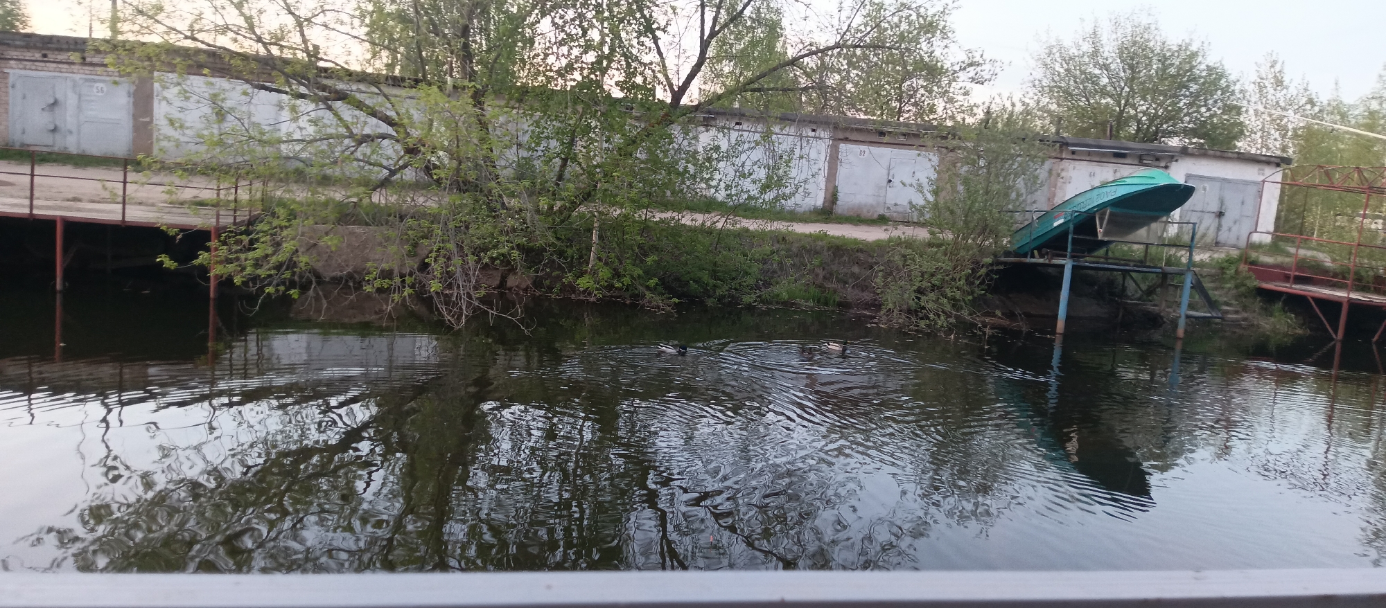 A scenic view near a body of water features a row of storage units/garages lining the backdrop. A small boat is positioned on a dock-like structure, partially extending over the water. Vegetation is abundant, with trees and bushes growing along the water's edge. Several ducks can be seen swimming in the water, creating ripples on the surface. Reflections of the surroundings are visible in the water. The foreground shows part of a railing or ledge, suggesting the vantage point from which the photograph was taken. The location appears to be a combination of industrial and natural elements. The city and region are both described as unknown.