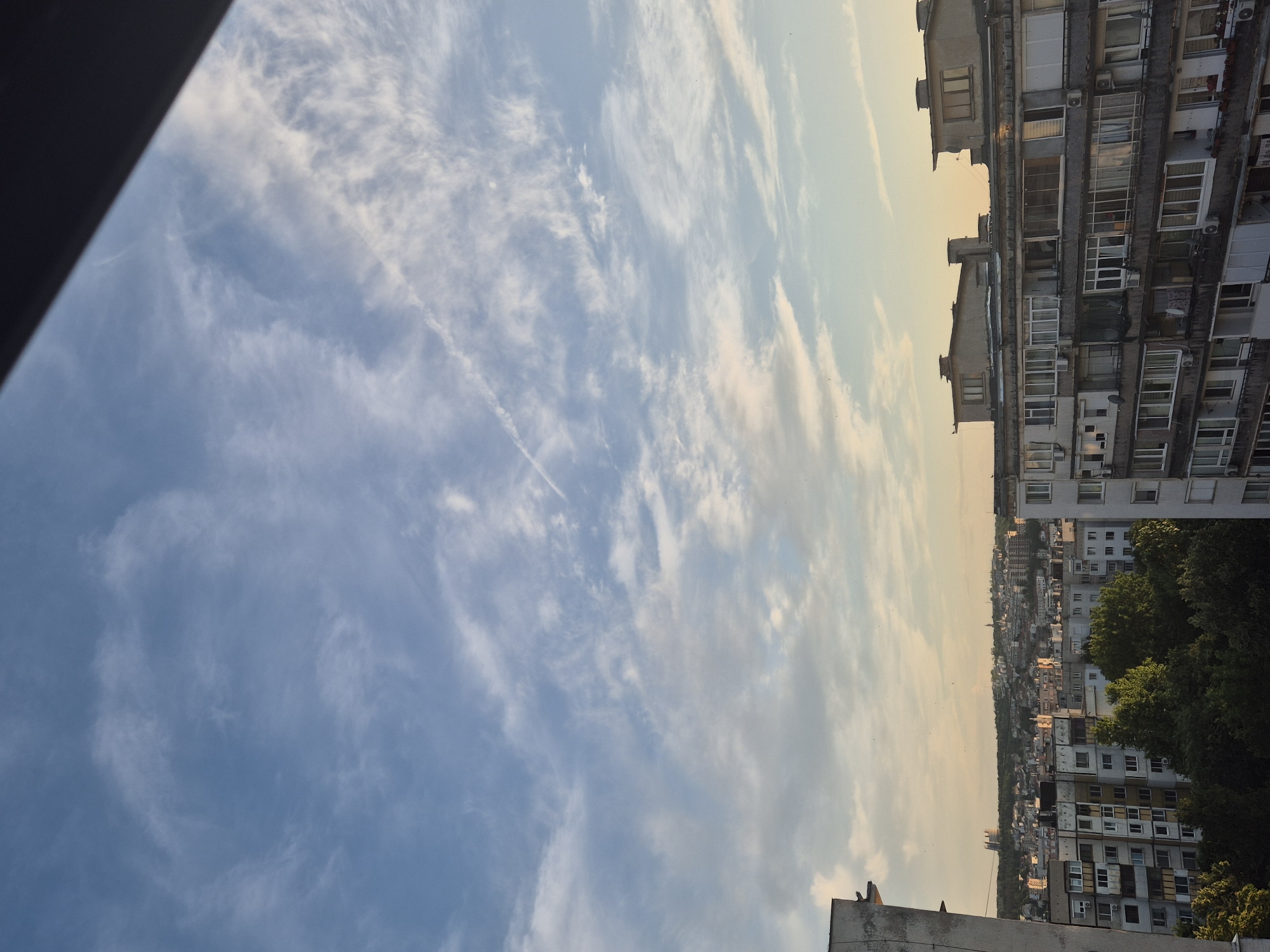 The image shows a view of a city skyline from a high vantage point, likely a balcony or window. The sky is a prominent feature, filled with wispy white clouds against a pale blue backdrop, with hints of sunset colors near the horizon. Below the sky, a dense urban landscape unfolds, characterized by rows of apartment buildings. These buildings appear to be of a similar architectural style, with repetitive window patterns and balconies. Some trees, dense and green, are visible between the buildings, suggesting a park or residential area with landscaping. In the distance, a larger cityscape with more varied structures and a hill can be seen. The lighting suggests it is either early morning or late afternoon, with soft, warm light illuminating parts of the city. A dark, angled shape occupies the top right corner of the frame, indicating the presence of an overhang or structure framing the view.
