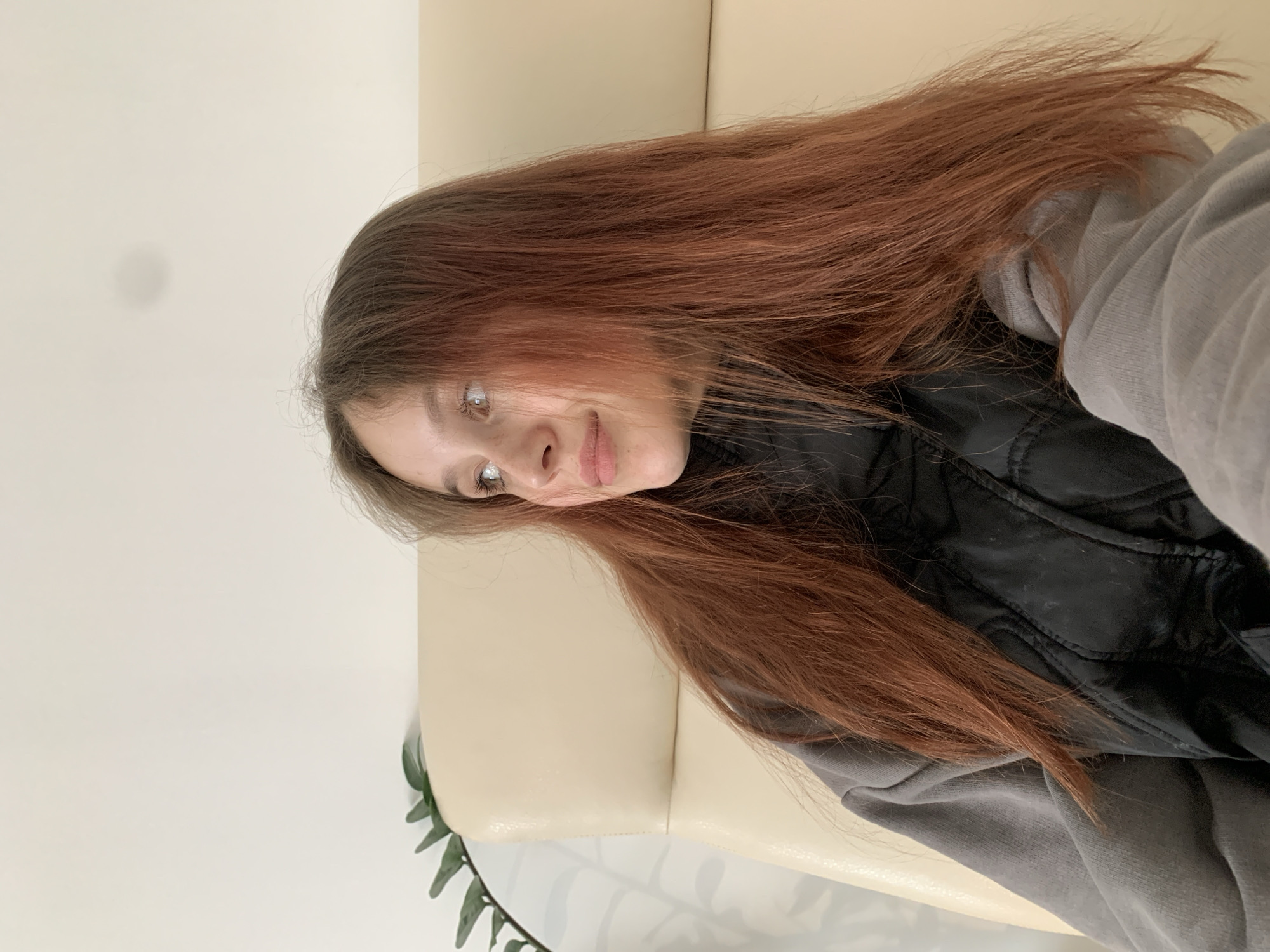 A woman with auburn hair is reclined on a beige sofa. She is wearing a grey hooded sweatshirt and a black vest. A green plant is visible in the lower left corner. The location in the city of Фоки, Russia, cannot be confirmed from the image.