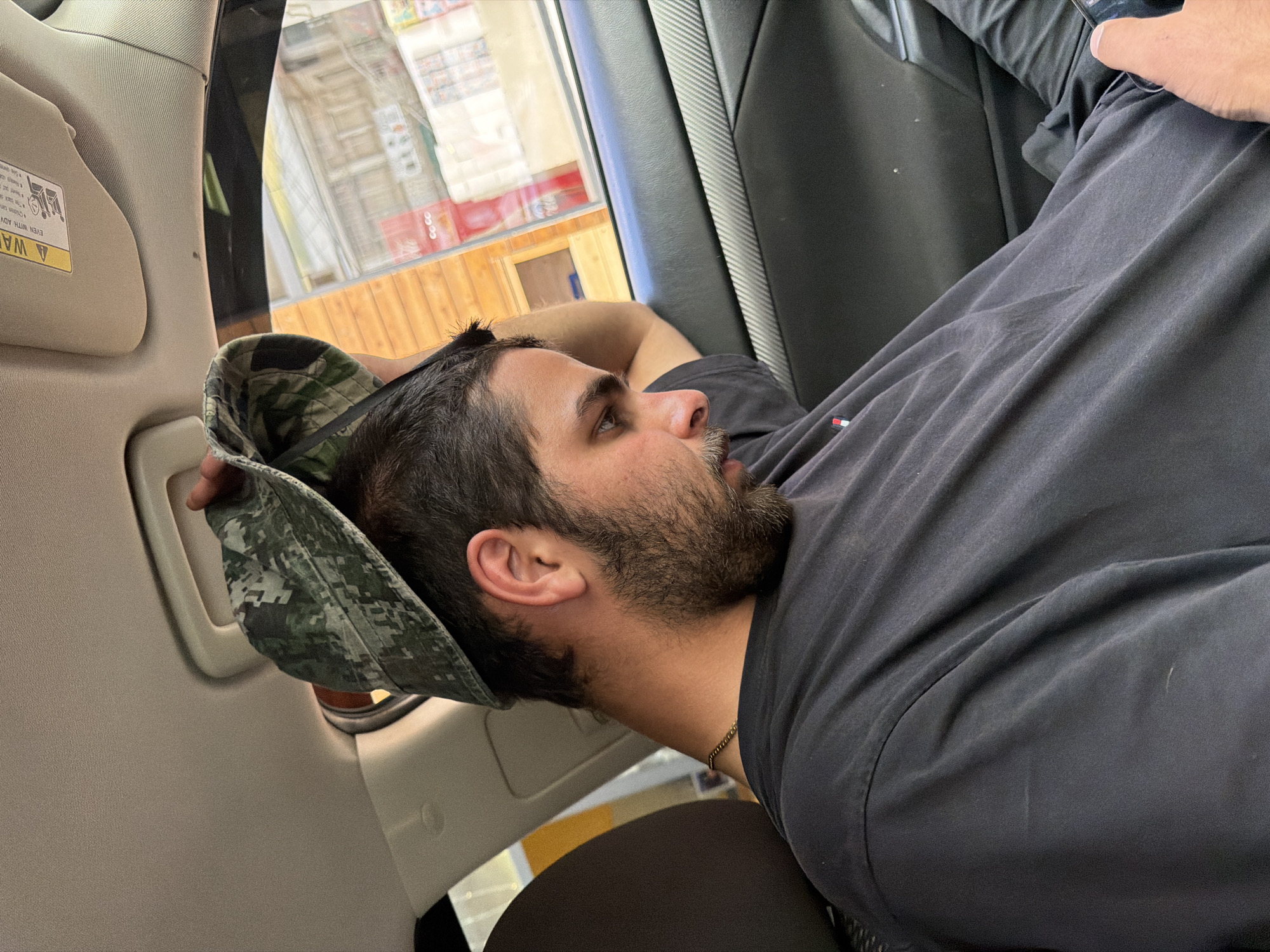 A young man with dark hair and a beard, wearing a black t-shirt and a camouflage bucket hat, is sitting in the passenger seat of a car. His head is tilted back and he appears to be resting his head against the window. His right arm is bent and resting on the interior of the car. The background shows a wooden building and storefronts through the car window. The interior of the car is dark, with a black dashboard and grey trim. The man is holding a phone in his right hand.